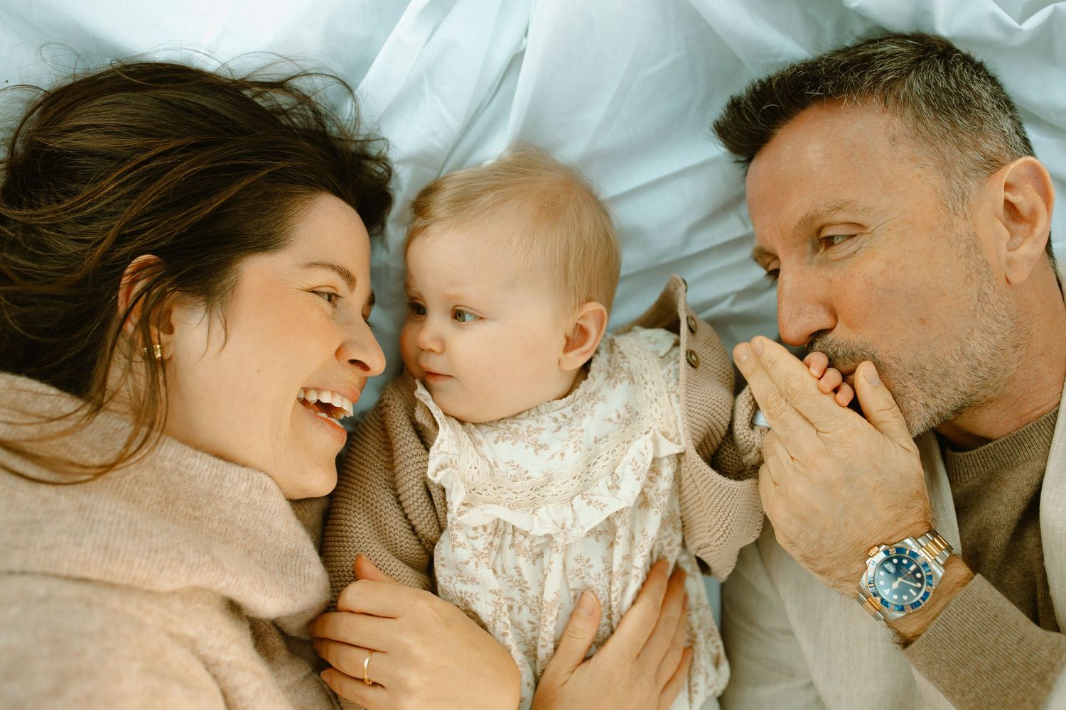 A family shares a joyful moment together while lying down, with their baby wearing a white dress between them.
