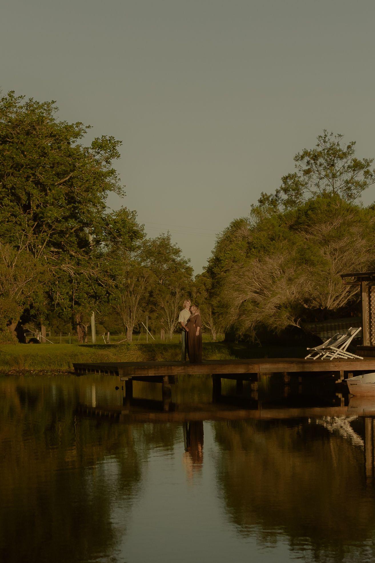 Tranquil lake scene at dusk with silhouetted trees and a wooden dock reflecting on still waters.