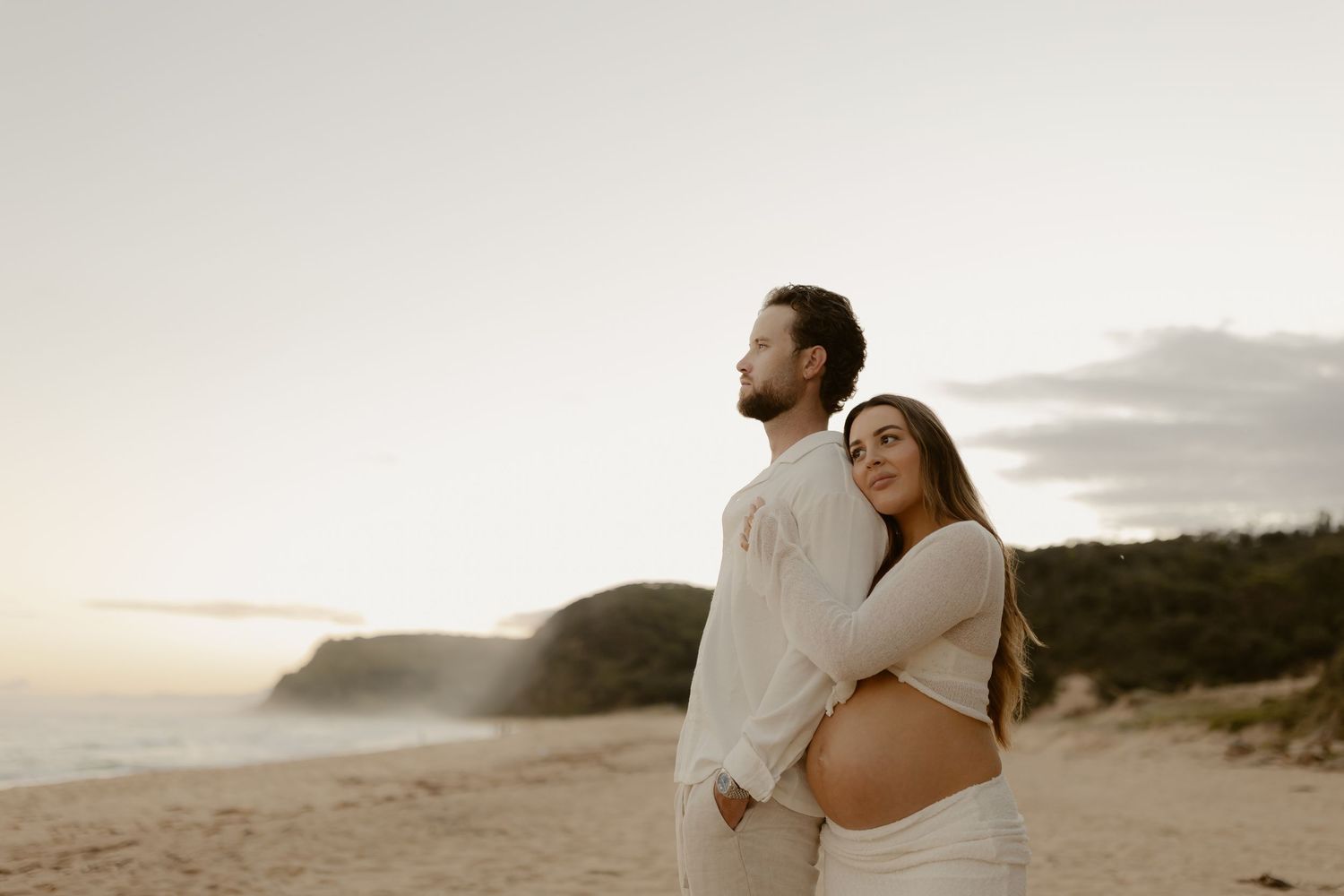 A couple embraces on a scenic beach at sunset with cliffs and coastline in the background.