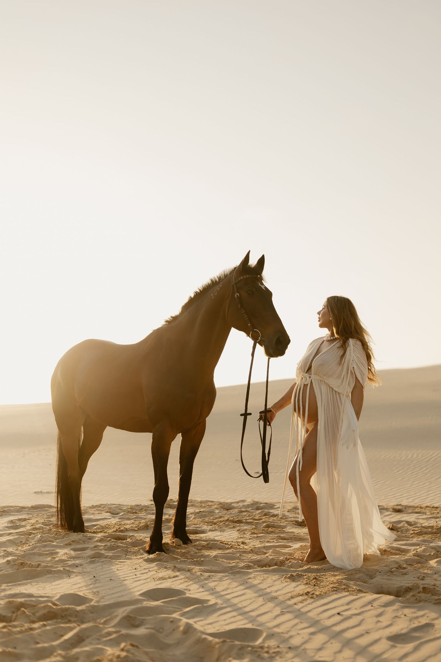 A horse and person in white dress create a dreamy scene on sandy dunes during golden hour.
