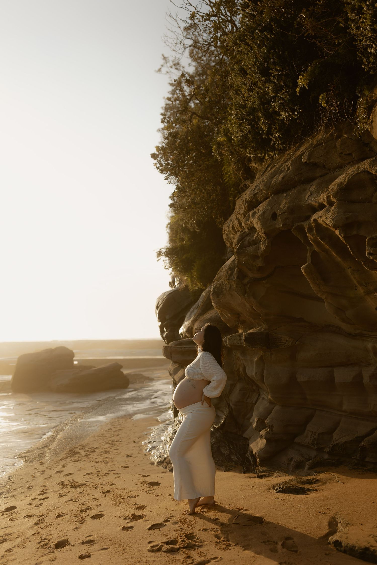 A person in white dress stands contemplatively near rocky cliffs at a misty beach during sunset.