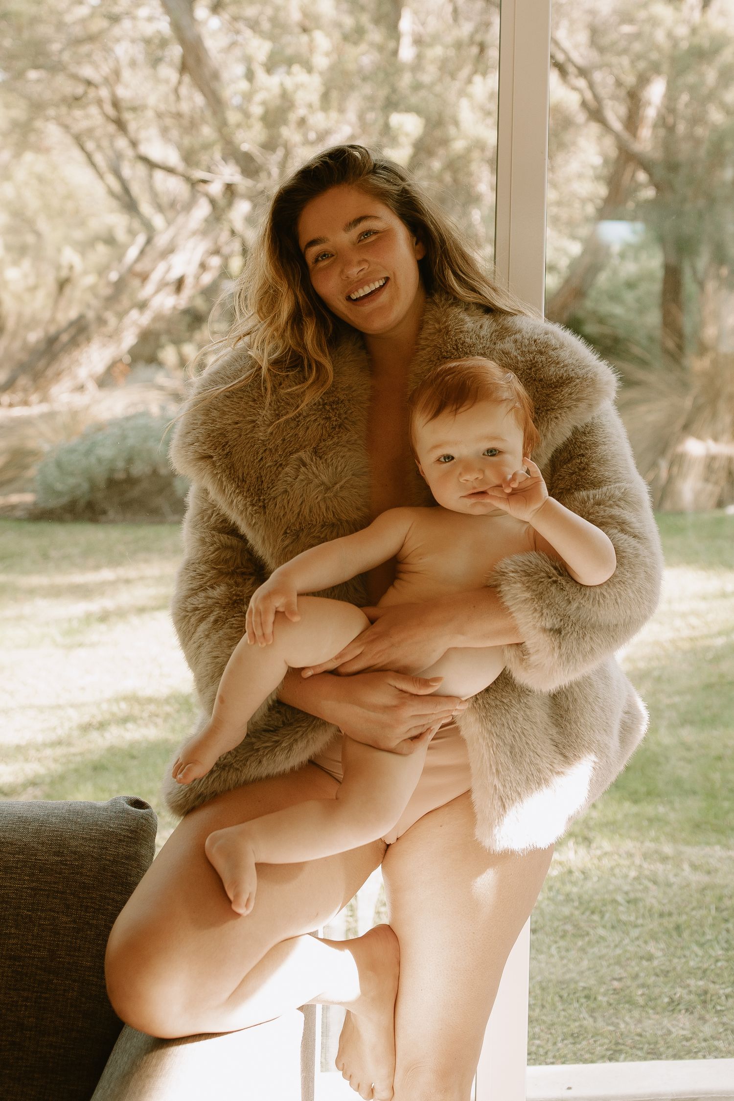 A tender moment captured outdoors as a cozy fur coat envelops parent and baby in warm, natural sunlight.