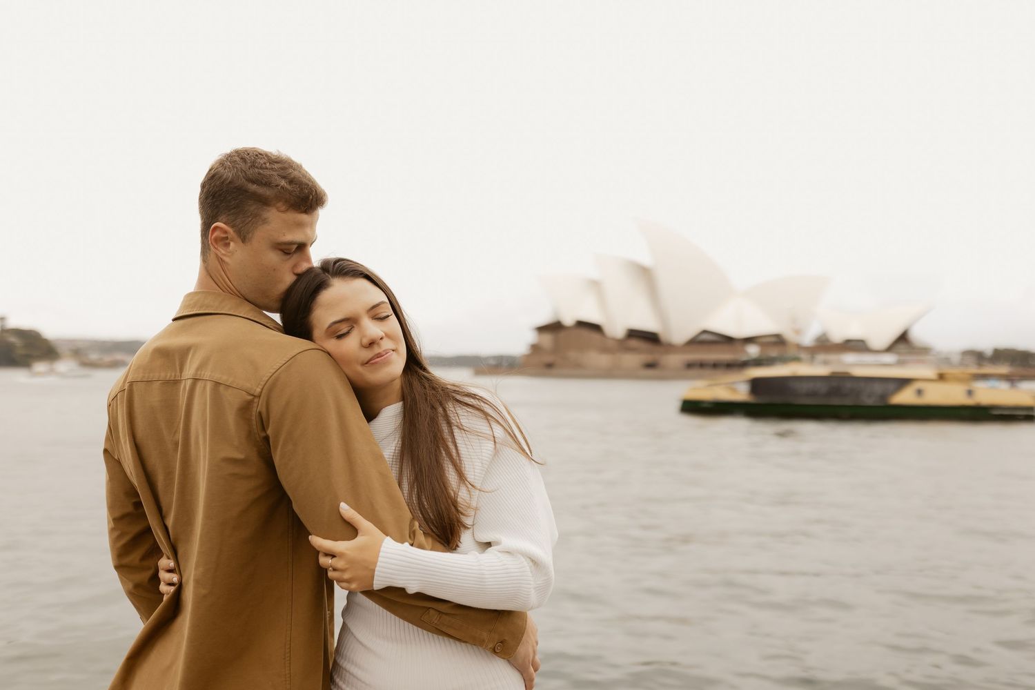A couple in warm clothing embracing by the harbour with the Sydney Opera House visible in the distance.