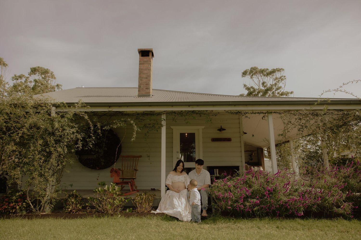Couple sitting together on porch of white cottage home surrounded by colorful flowering garden.