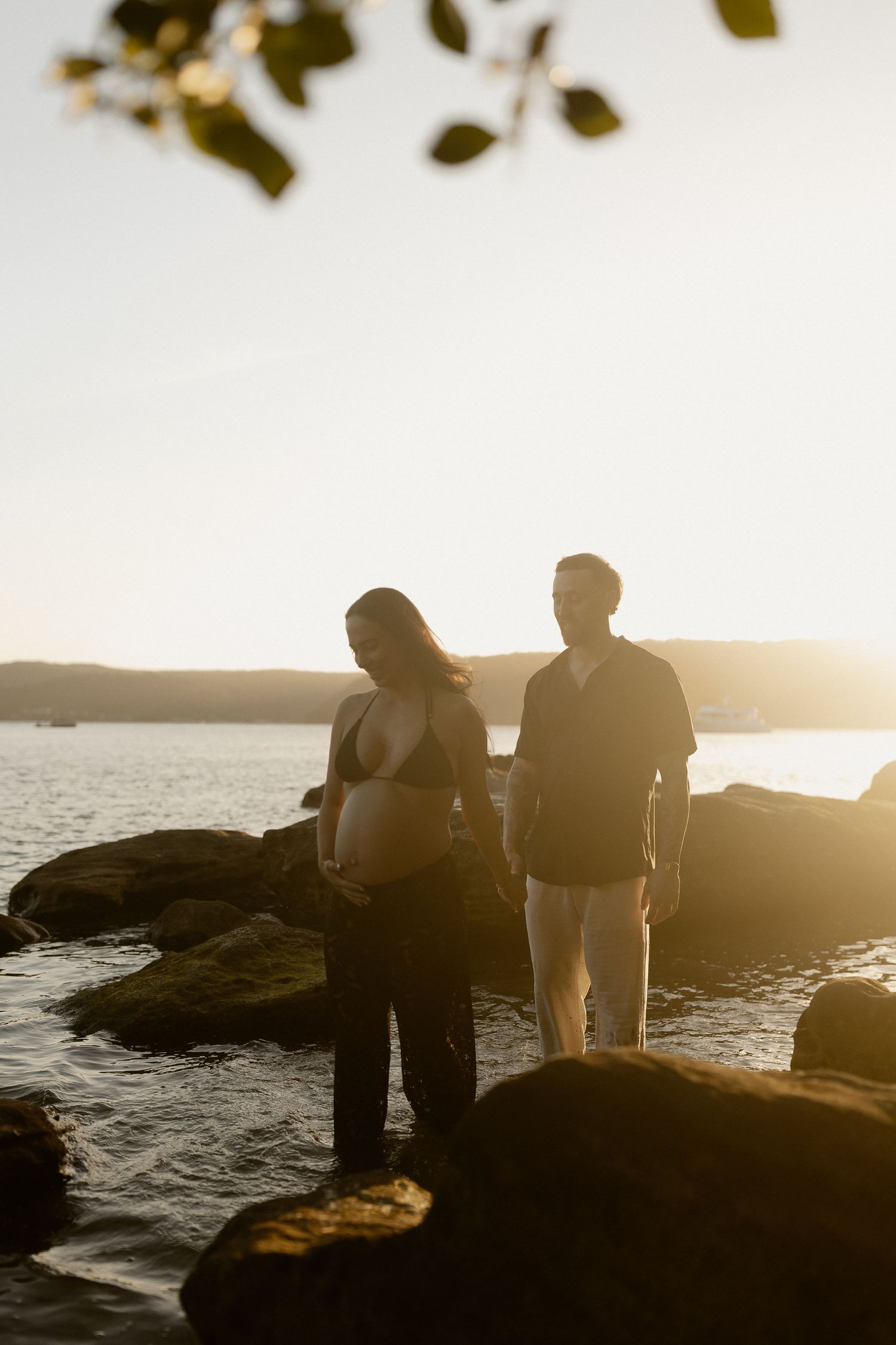 Two figures stand silhouetted against a sunset on rocky coastal shoreline as golden light illuminates the peaceful scene.
