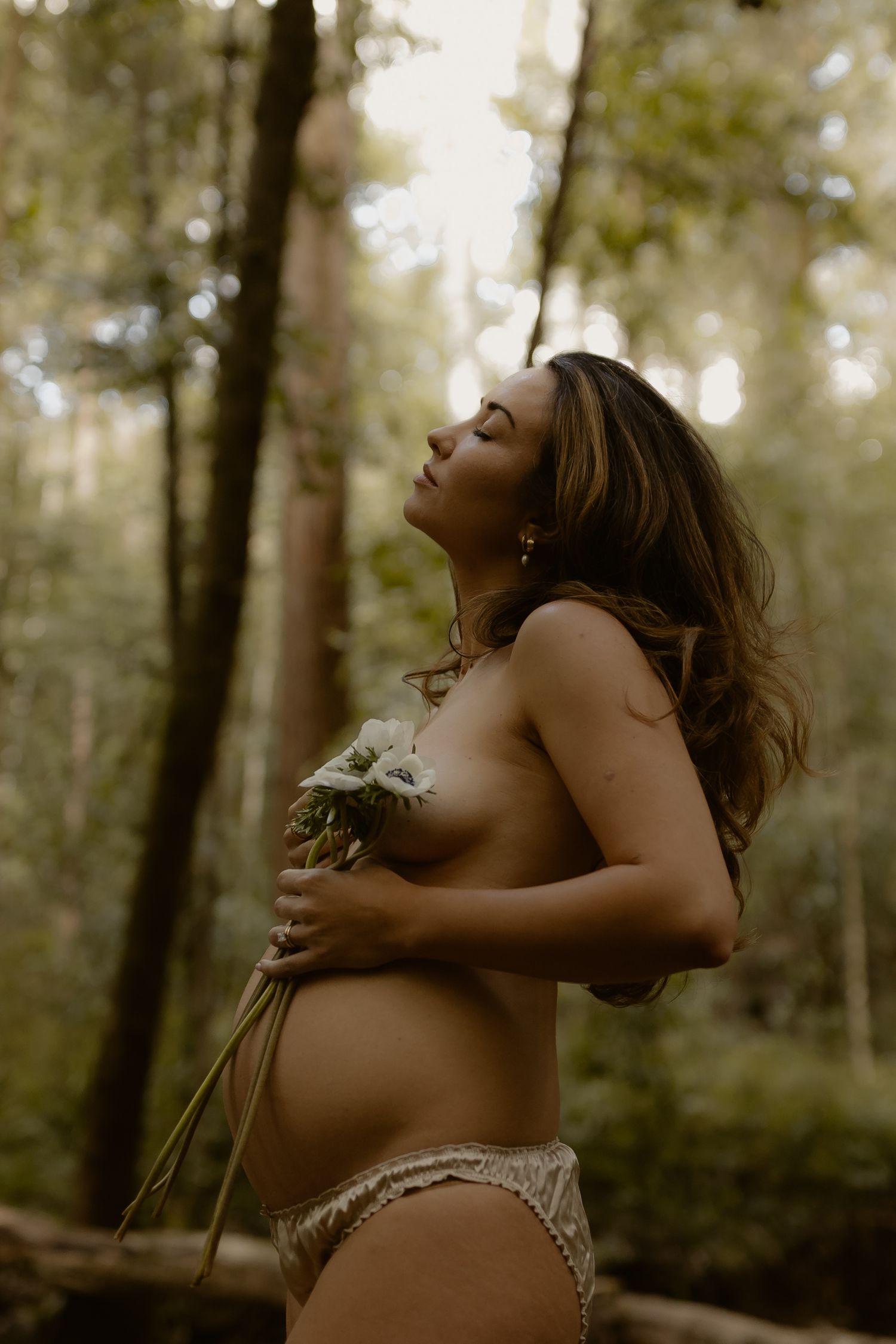 A figure in brown lace poses dramatically in a dreamy forest setting with dappled sunlight filtering through trees.