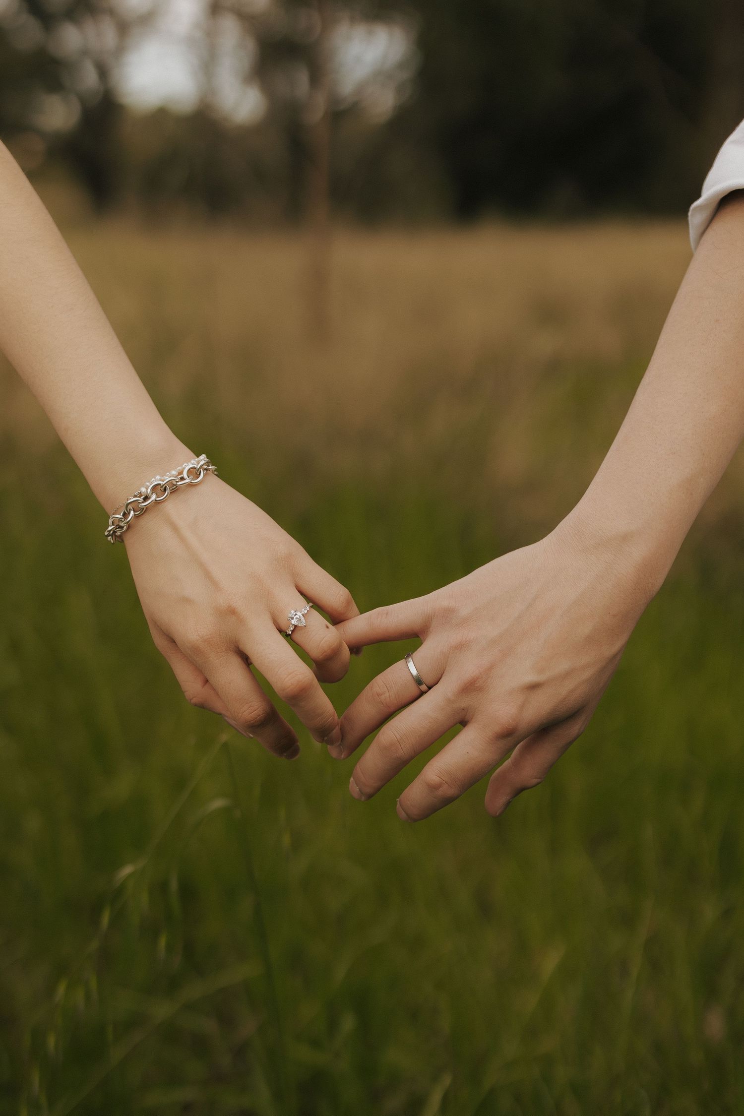 Close-up of hands wearing rings reaching towards each other in a grassy field during golden hour.