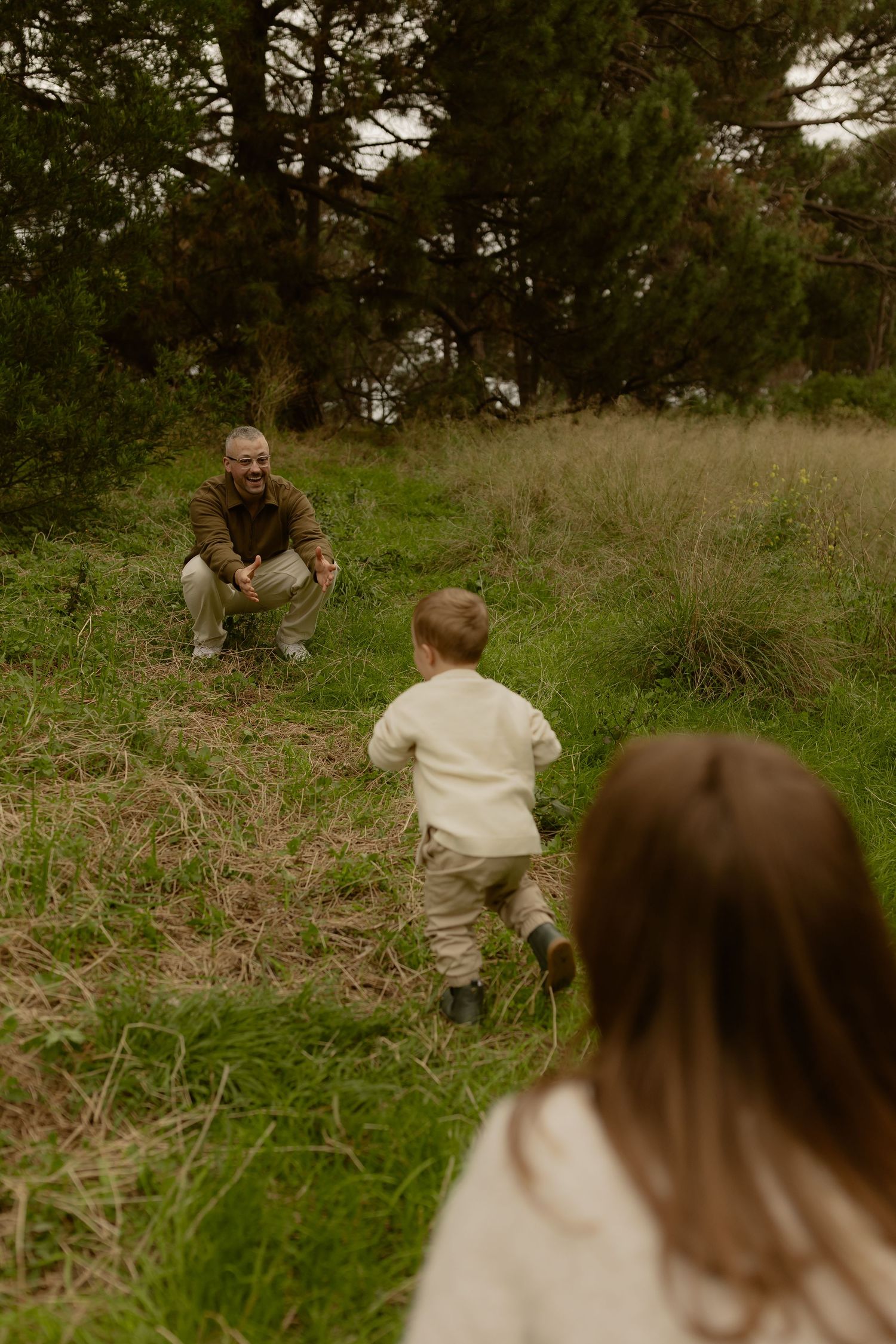 A toddler runs toward a crouching man on a grassy hillside during an outdoor family photoshoot.