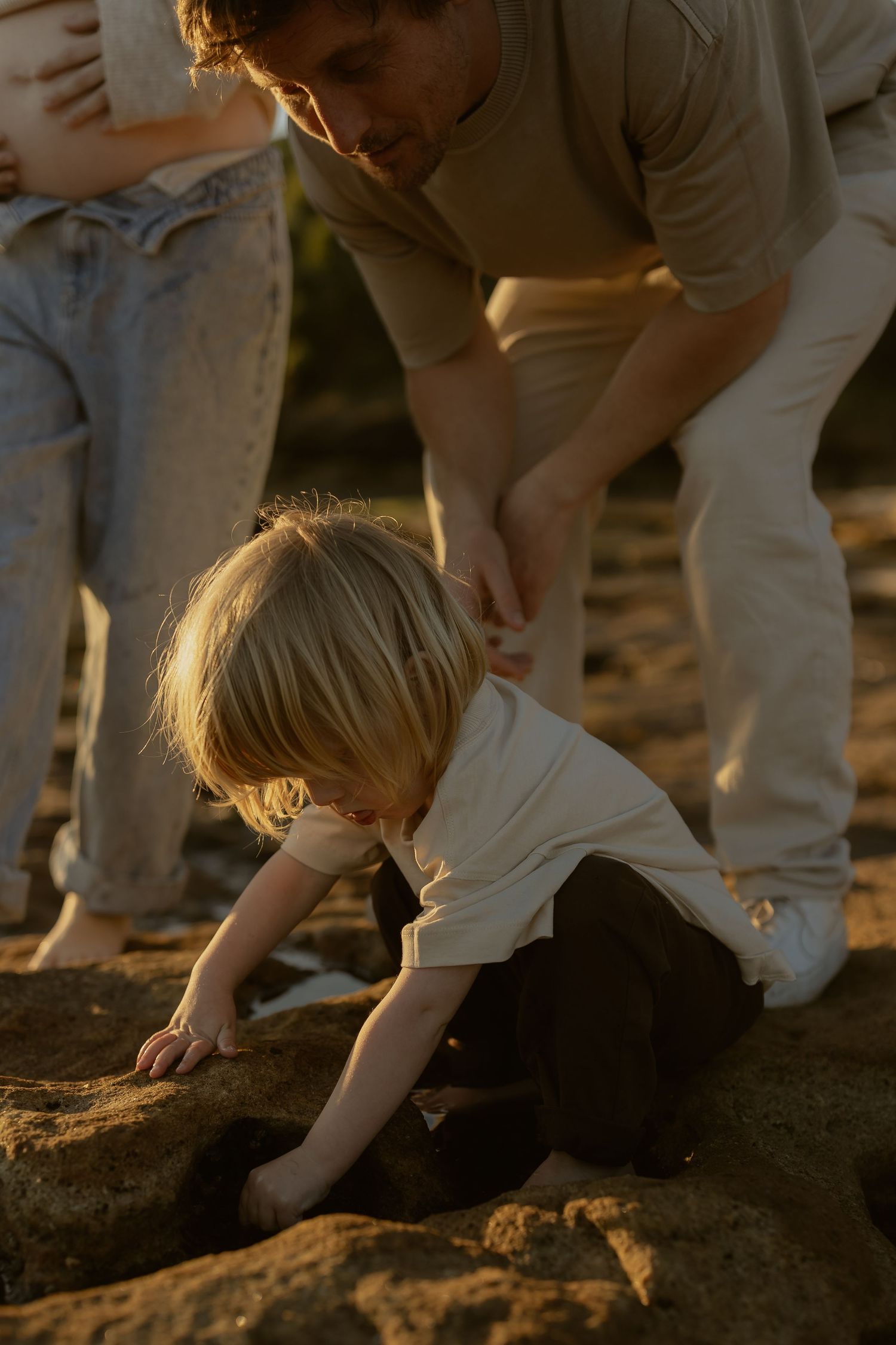 A young blonde child explores rocks on a beach while parents watch in warm golden sunset light.