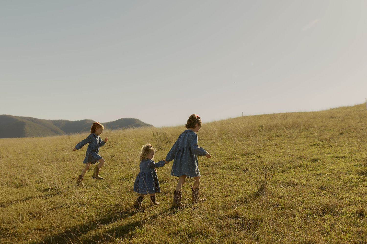 Three young girls in blue dresses holding hands and running through a sunny green hillside meadow.
