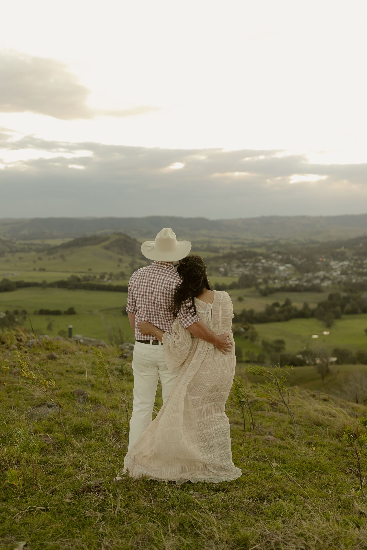 Couple embracing on a hilltop at sunset, overlooking a lush green valley, man in cowboy hat and woman in white dress.