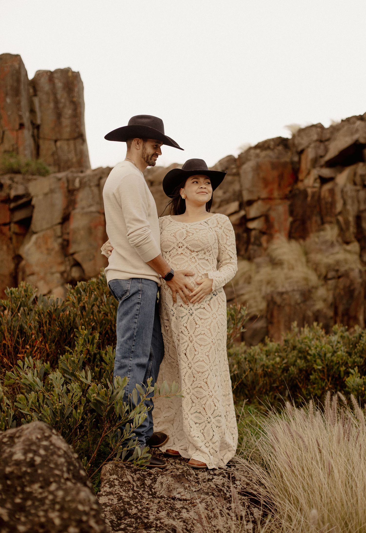 A couple in western attire shares a romantic moment against rugged desert cliffs at sunset.