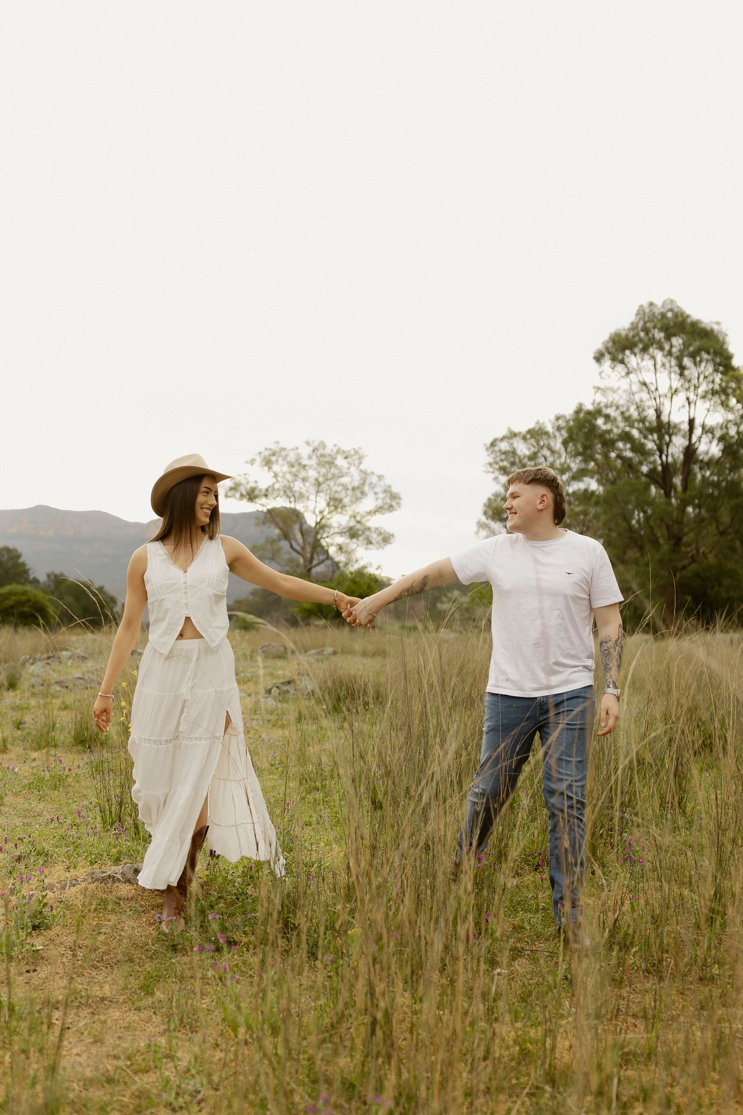A couple holds hands while standing in tall grass with mountains in the distance.
