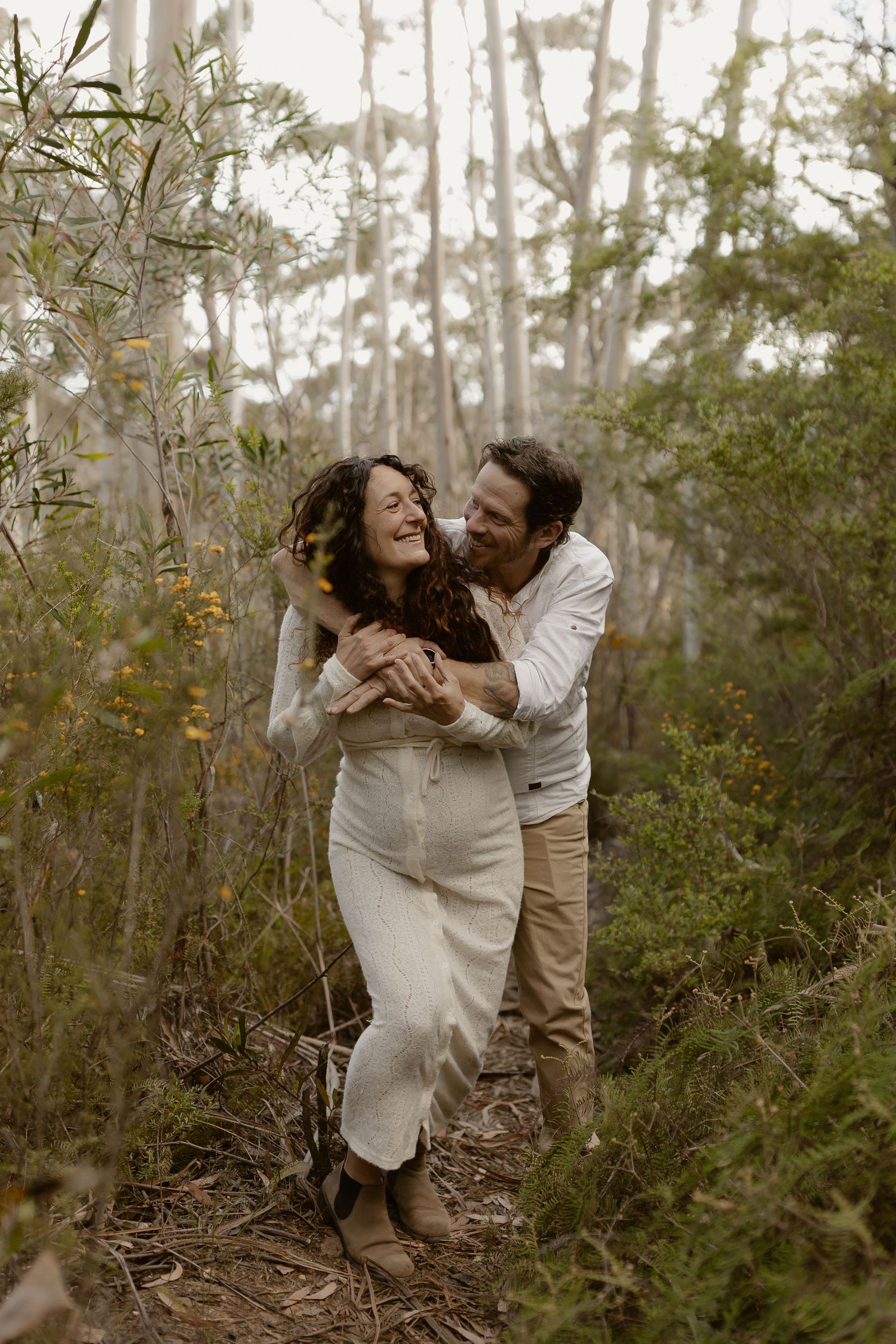 A romantic couple embraces playfully among trees and foliage in a natural outdoor setting.
