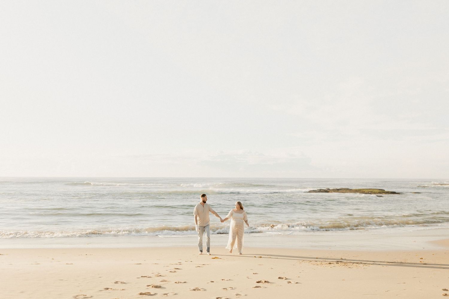 A couple walks along a misty beach shoreline while holding hands on an overcast day.