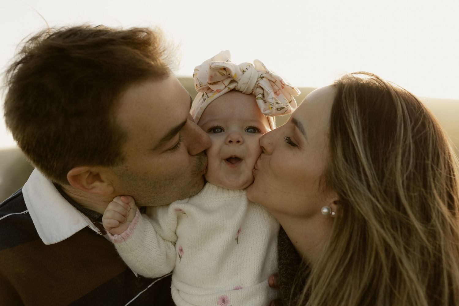 Parents kissing their smiling baby girl wearing a pink bow headband in a warm, golden-lit family portrait.