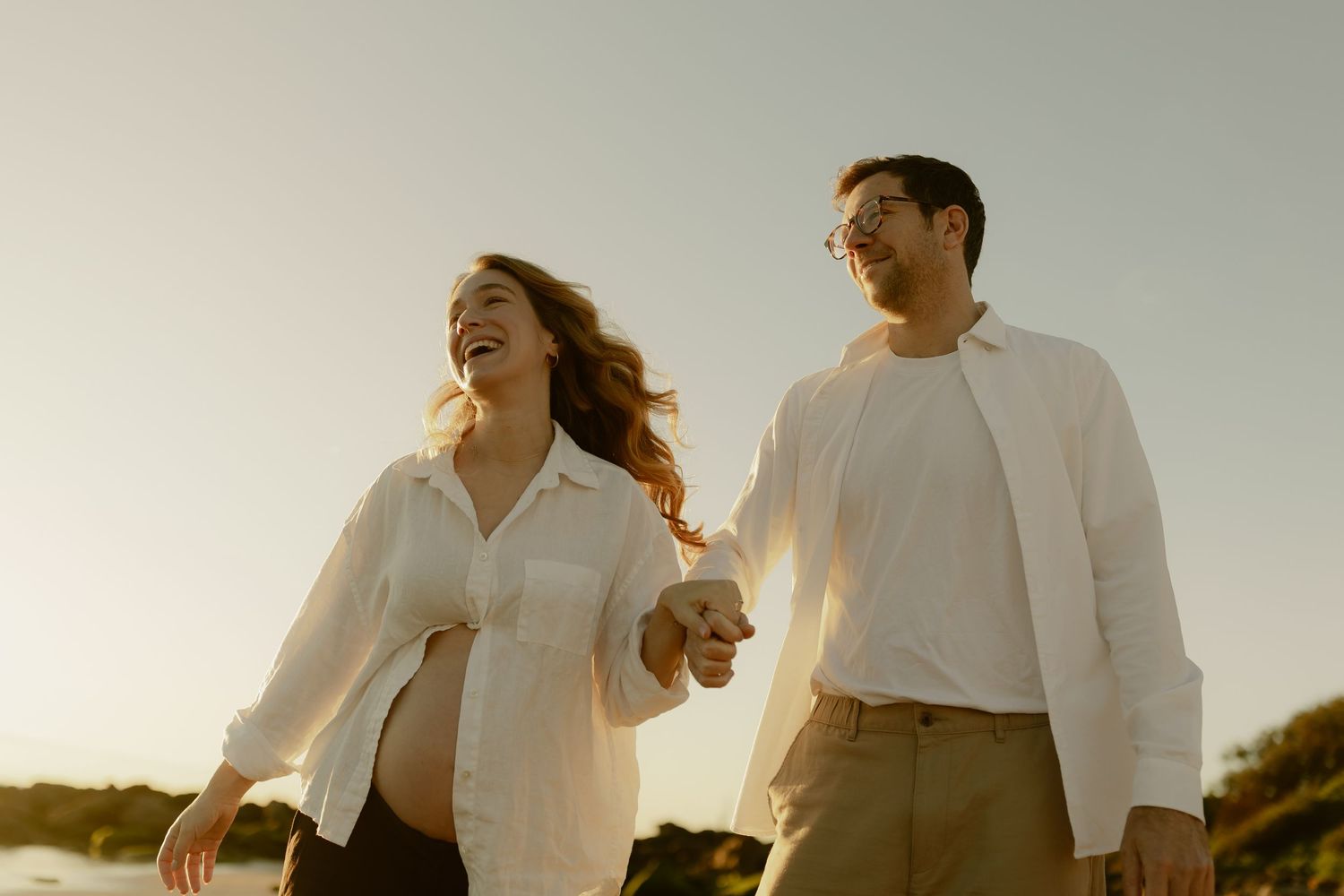 Pregnant couple holding hands laughing on a rocky beach at golden hour, woman in white shirt, man in glasses.