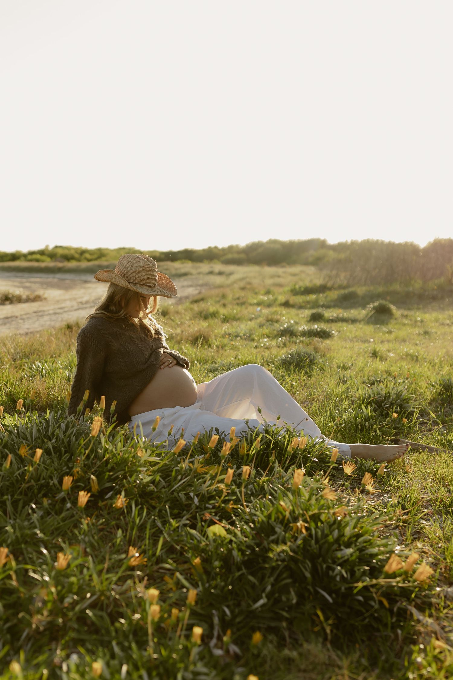 Pregnant woman in straw hat and white pants sitting in green field with yellow wildflowers at golden hour.