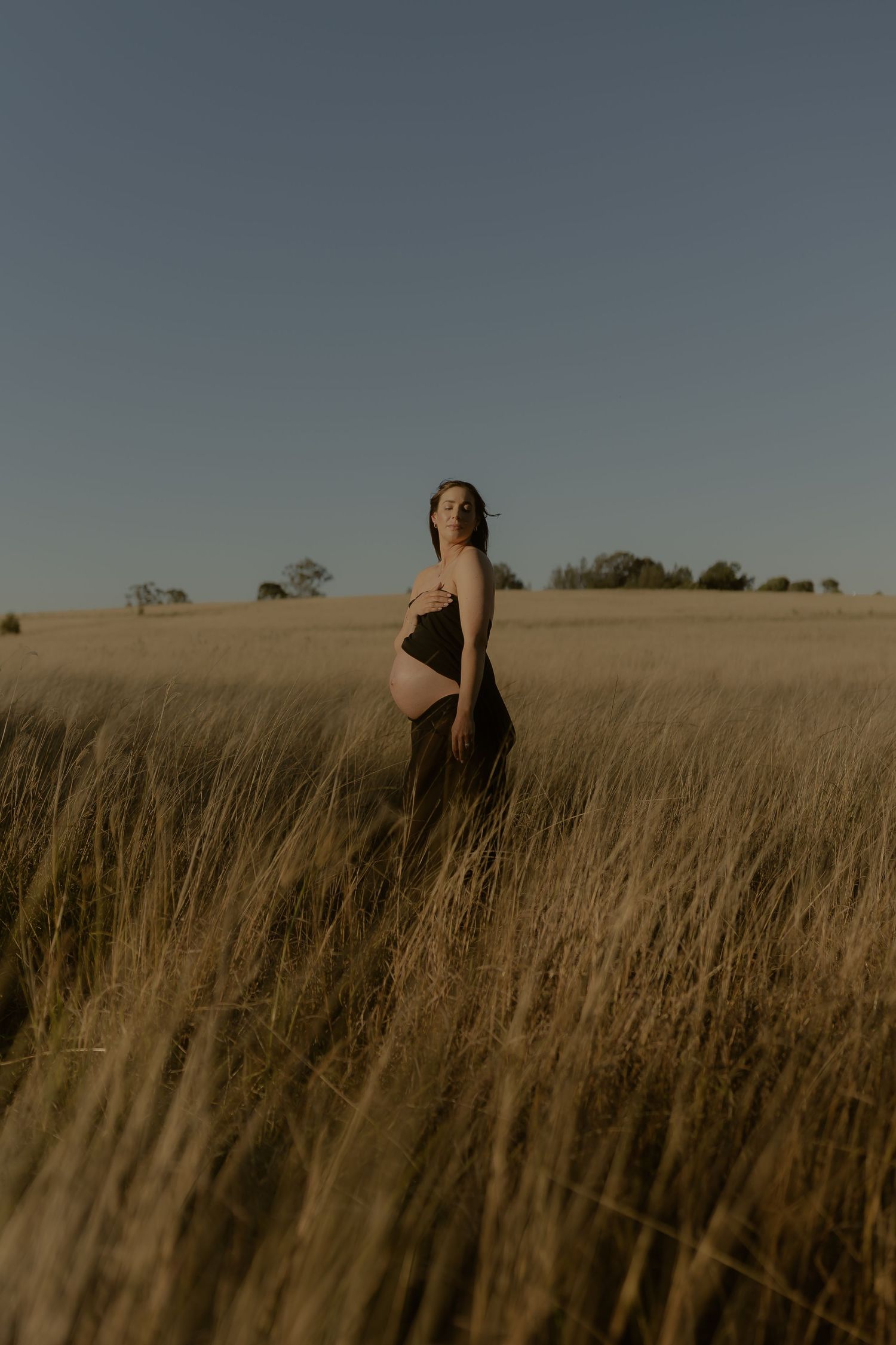 Pregnant woman standing in golden grass field wearing dark outfit under clear blue sky.