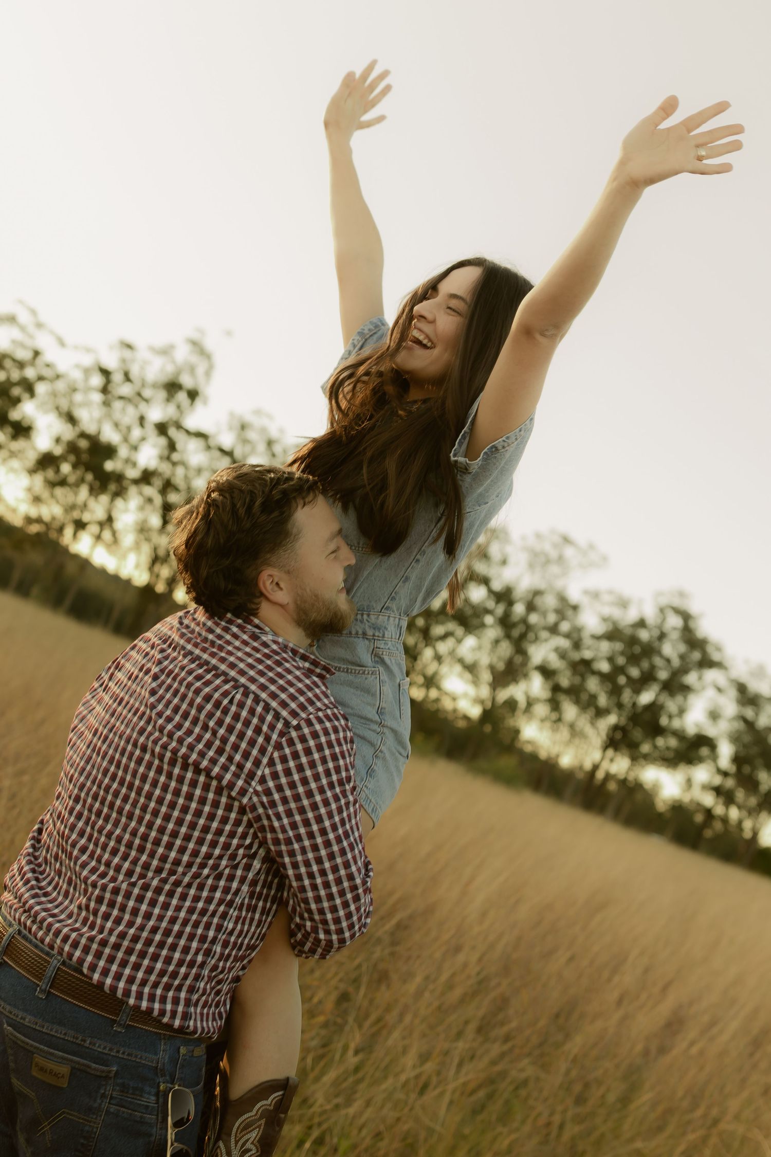 A joyful woman raises her arms while being lifted by a man in a sunlit grassy field.