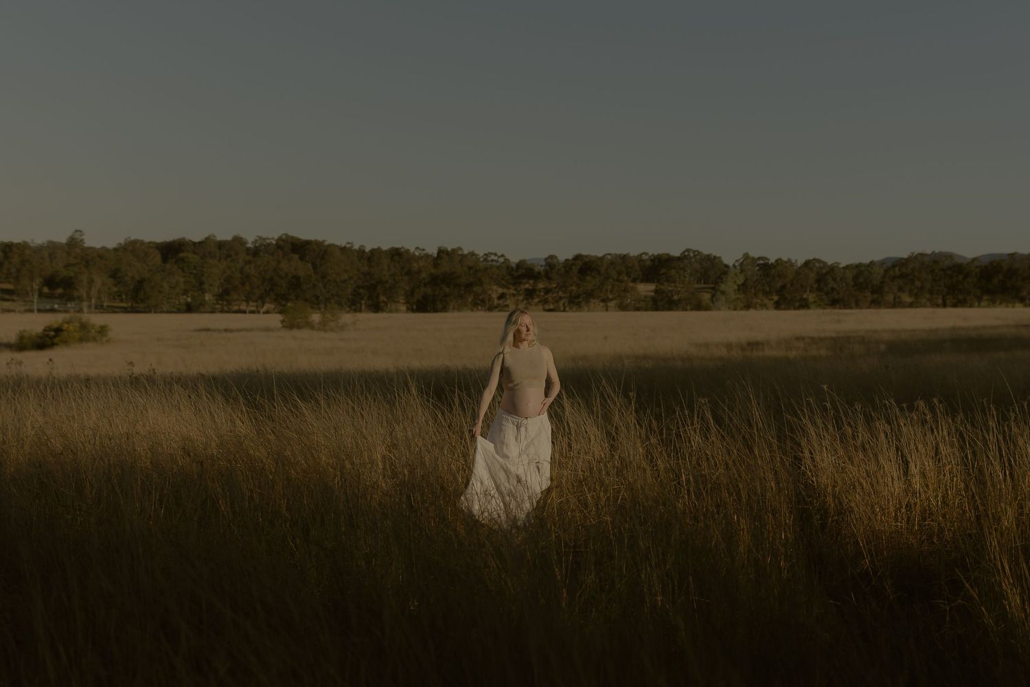 A woman in a white dress stands in tall golden grass fields with trees and clear sky in the background.