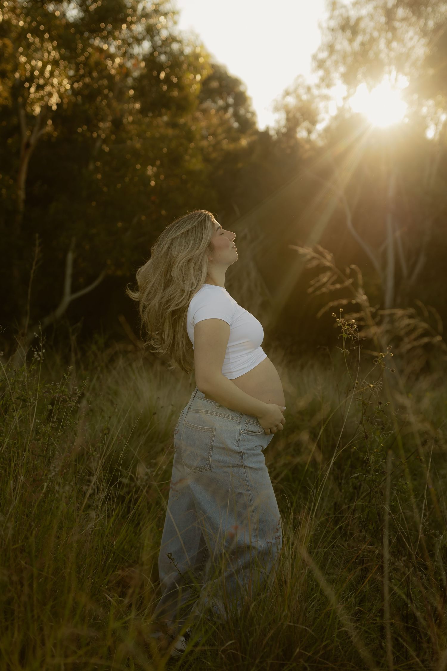 Pregnant woman in white crop top and denim skirt standing in sunlit meadow, cradling baby bump at golden hour.