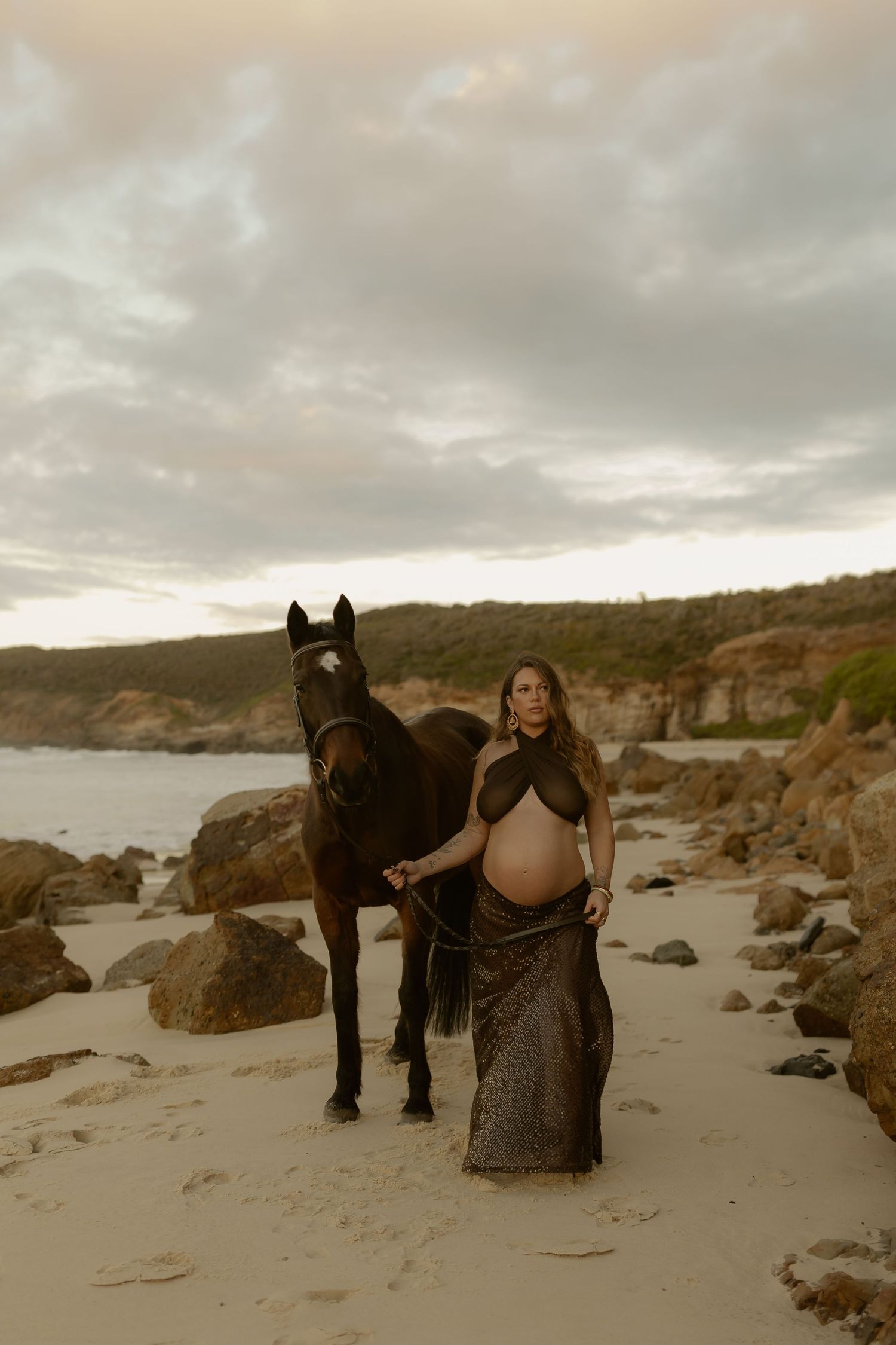 Pregnant woman in flowing dark wrap stands with brown horse on sandy beach at sunset with rocky coastline.