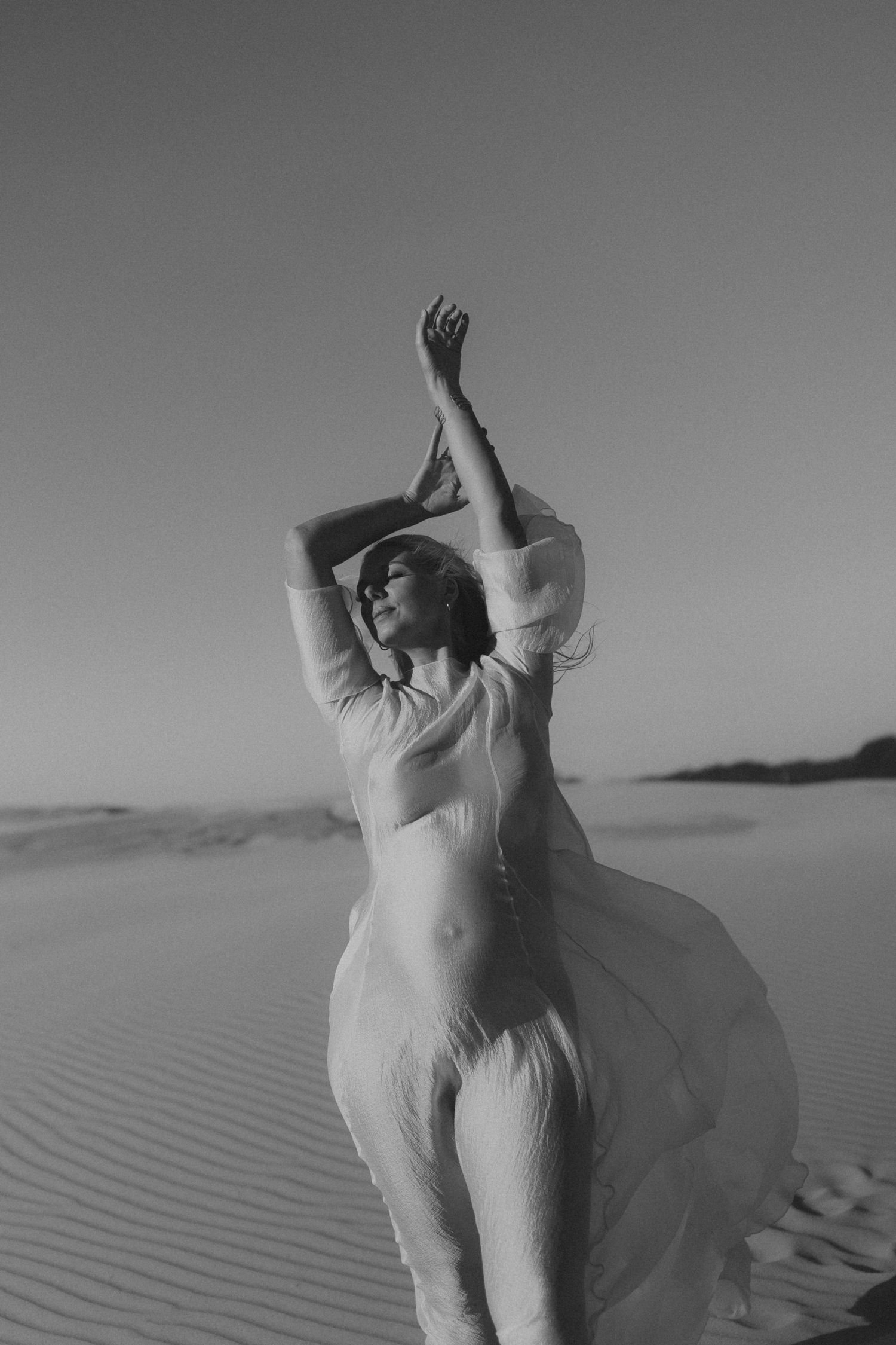Black and white photo of a woman in flowing sheer fabric posing gracefully on sand dunes with arms raised.