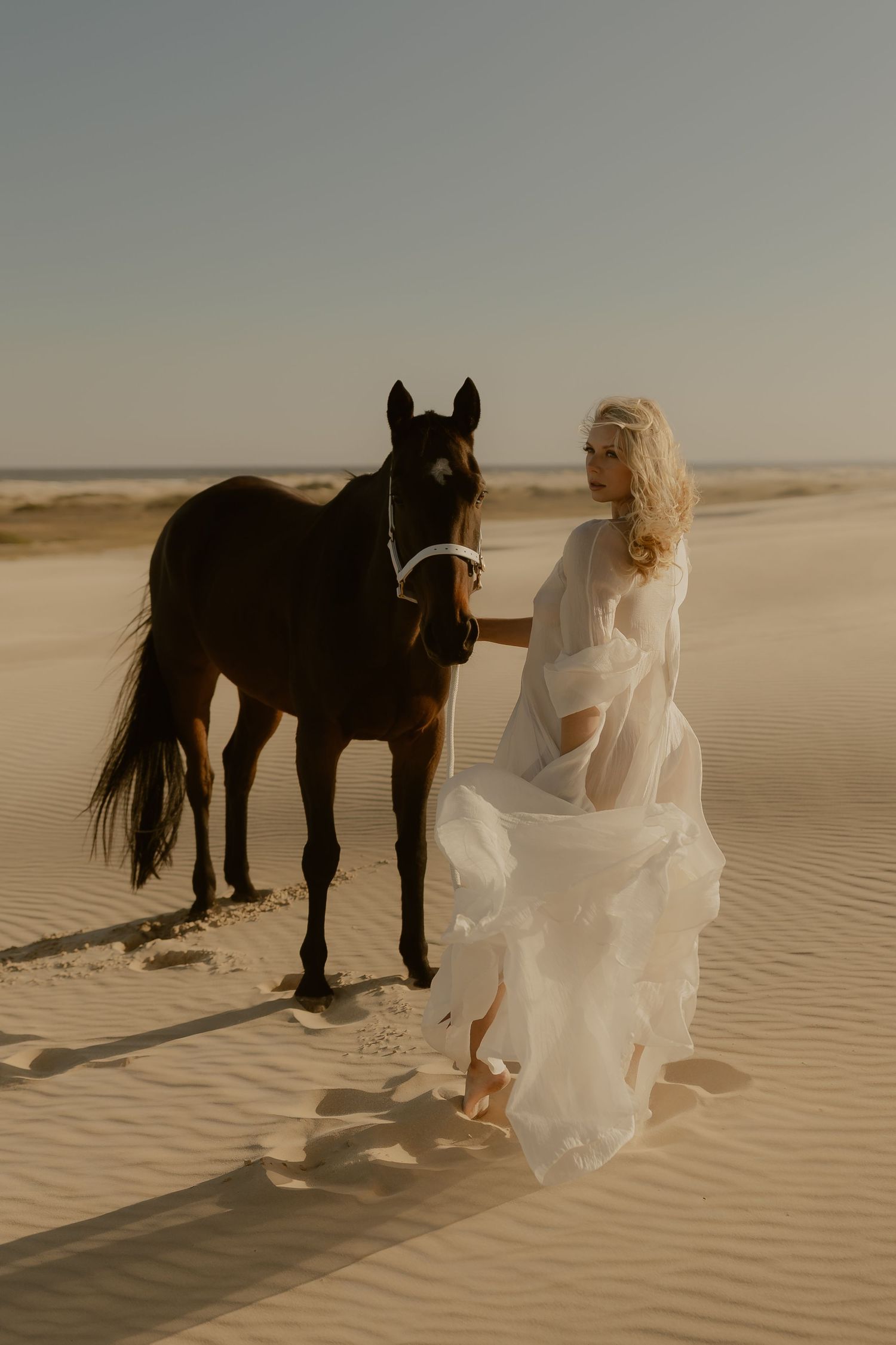 Blonde woman in flowing white dress walking with a dark brown horse on sandy desert dunes.