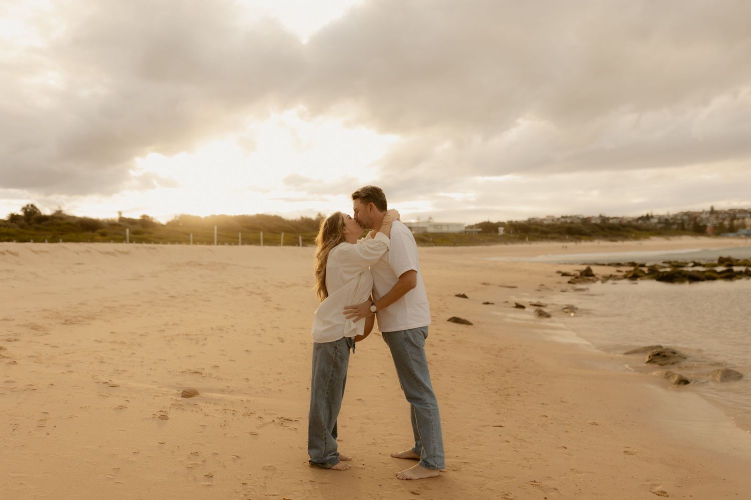Couple kissing on a sandy beach at golden sunset, wearing casual white shirts and jeans near the ocean shoreline.