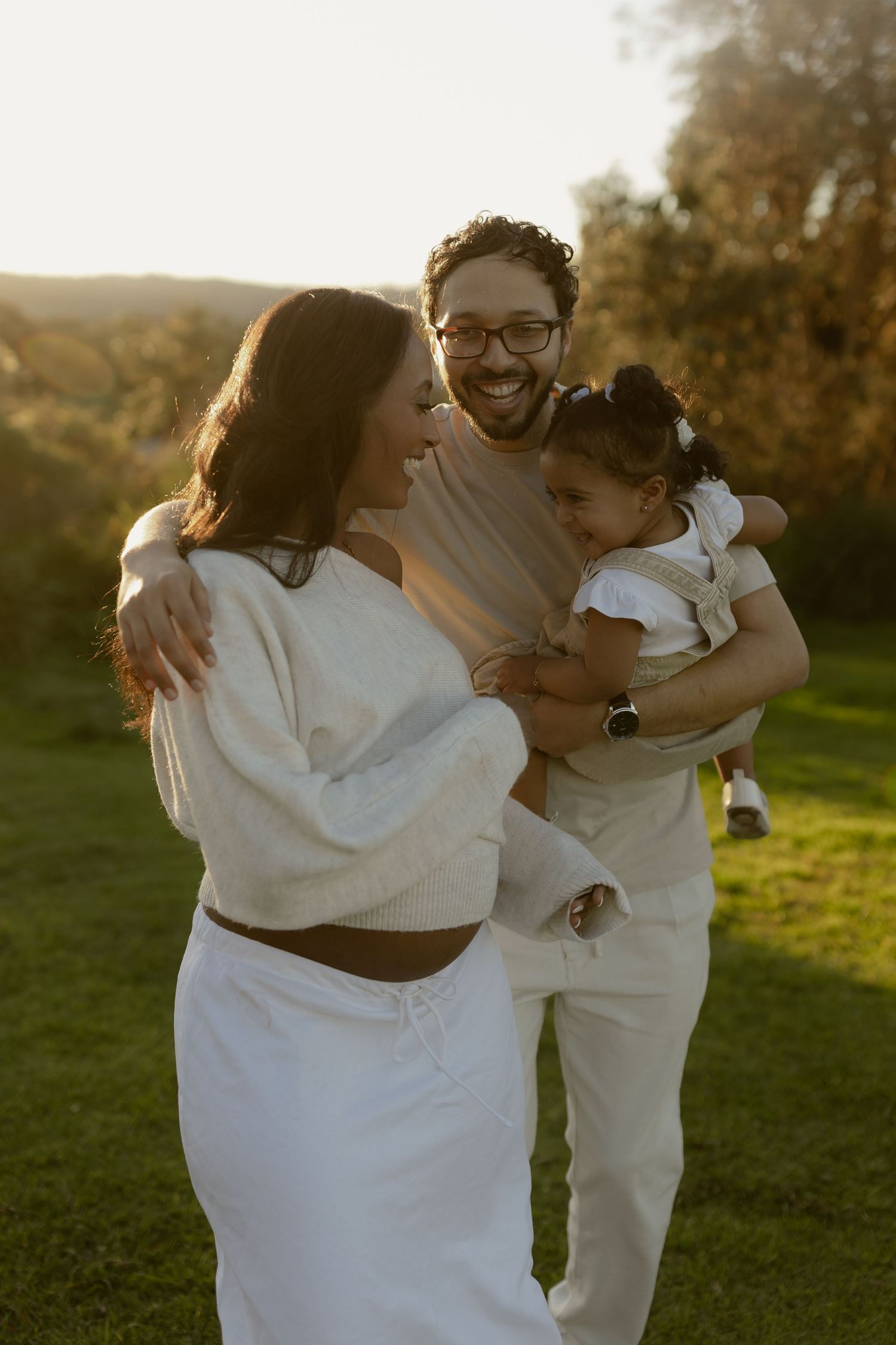 Happy family with father holding toddler daughter and mother laughing together in golden sunset light.