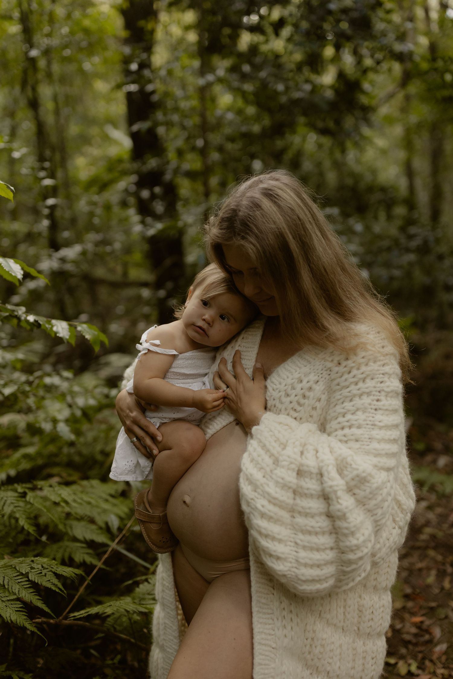 Pregnant woman in white cardigan holds toddler in lush green forest setting.