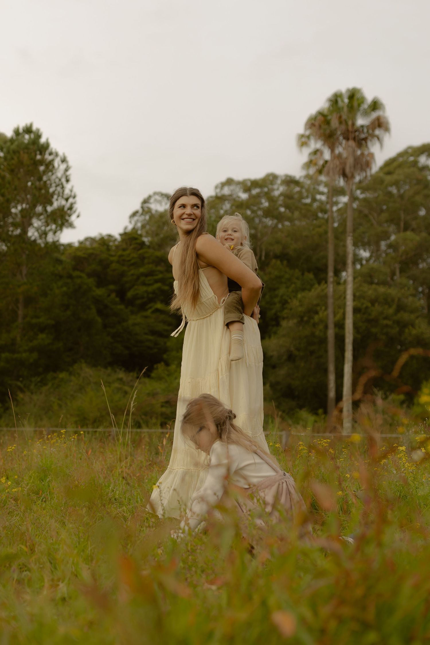 A smiling woman in a cream dress holds a baby while a child plays in a wildflower field.