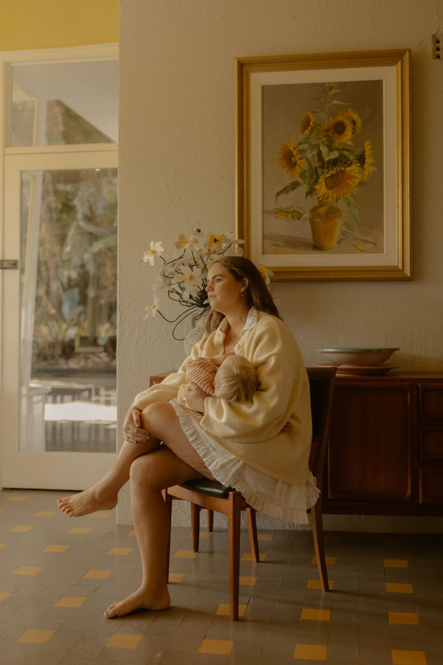 Mother in yellow robe breastfeeding baby on chair beside sunflower painting in warm vintage interior.