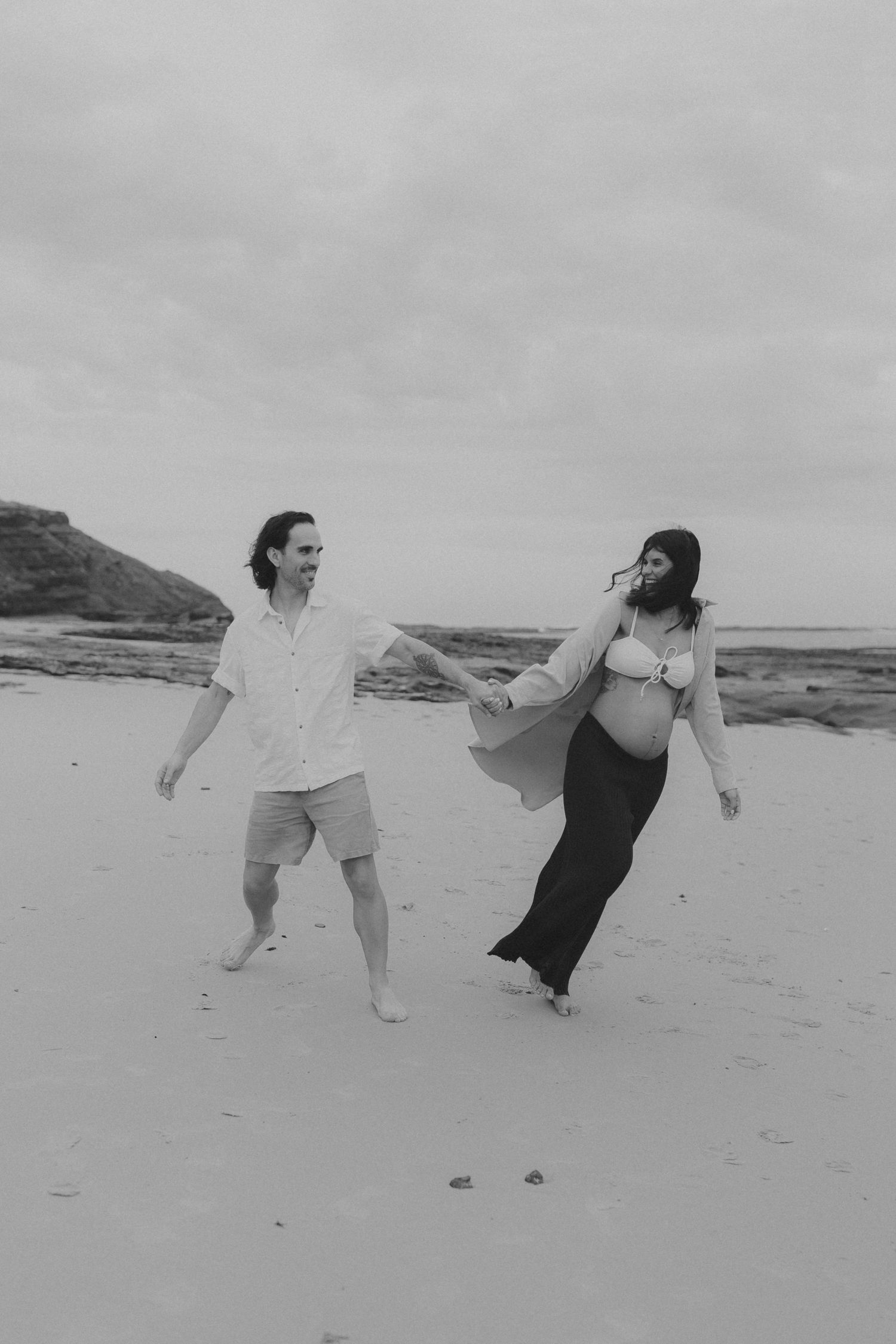 A black and white photo of a couple dancing joyfully together on a beach.