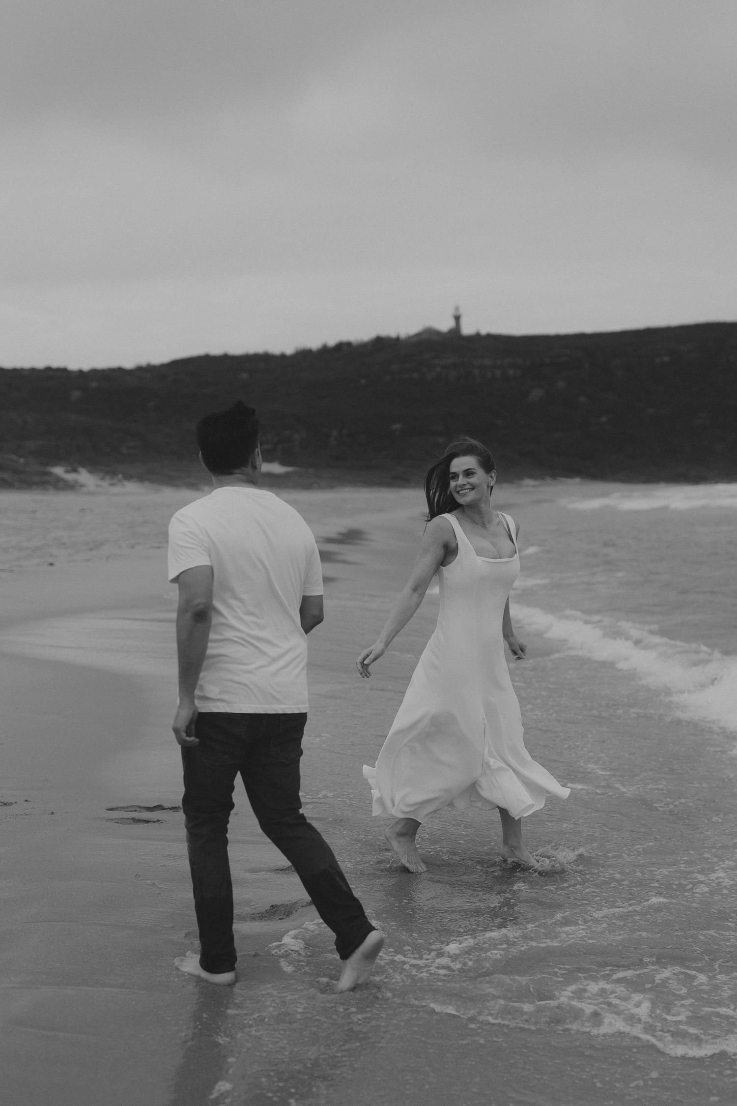 A couple plays in the ocean waves, captured in black and white photography.
