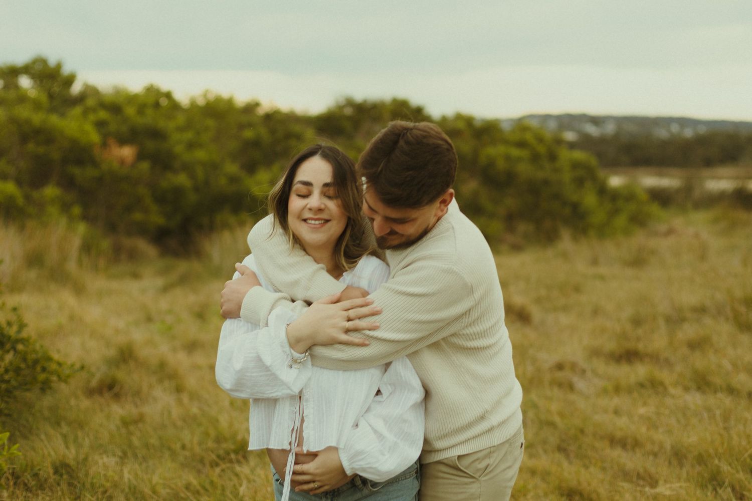 A couple embraces and laughs together in a golden sunlit field with green hills in the background.
