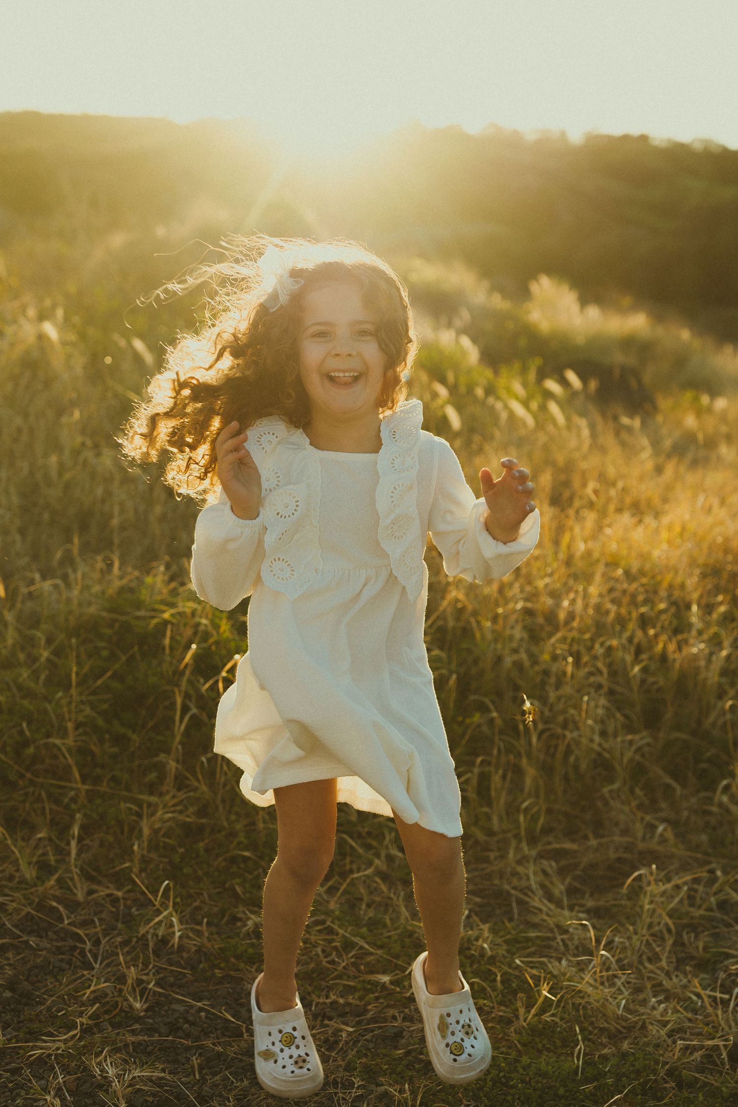 Young child joyfully jumping and playing in a sun-drenched field wearing a white dress.