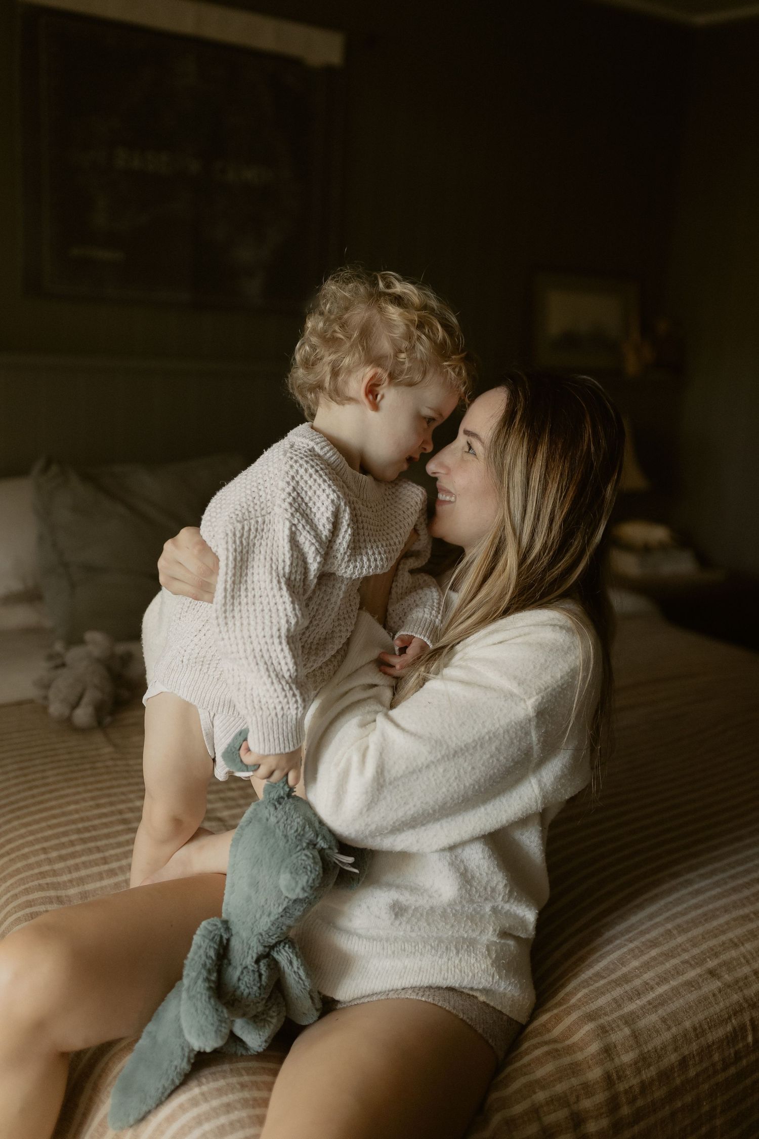 Intimate indoor portrait showing tender moment between parent and child in cozy white sweaters on a bed.