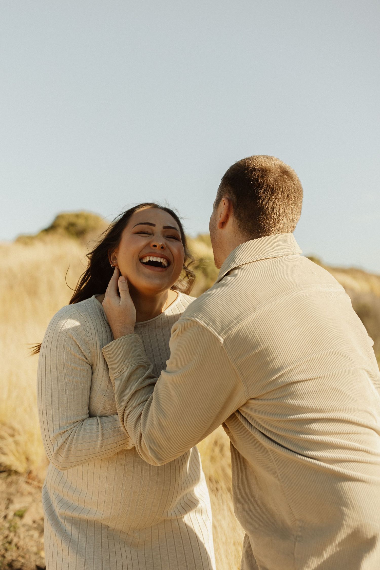 Intimate moment between couple laughing together in warm sunlight against a natural outdoor backdrop.