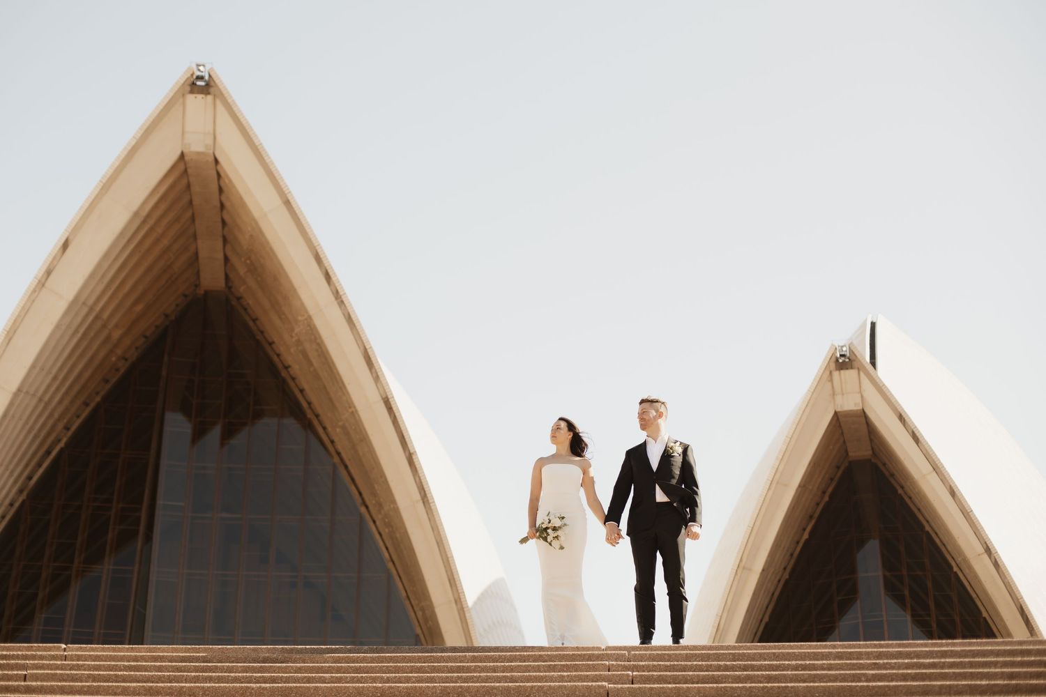 A couple in wedding attire stands between the iconic pointed peaks of the Sydney Opera House in Australia.