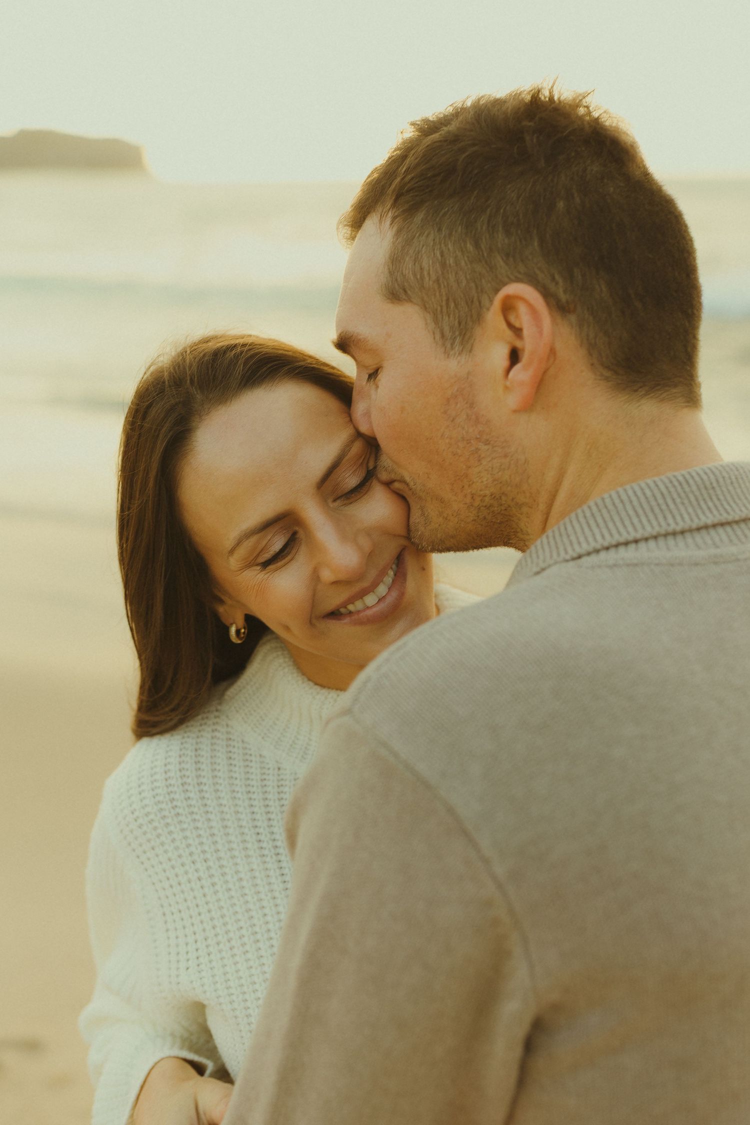 A romantic couple shares an intimate moment at sunset on the beach while wearing cozy sweaters.
