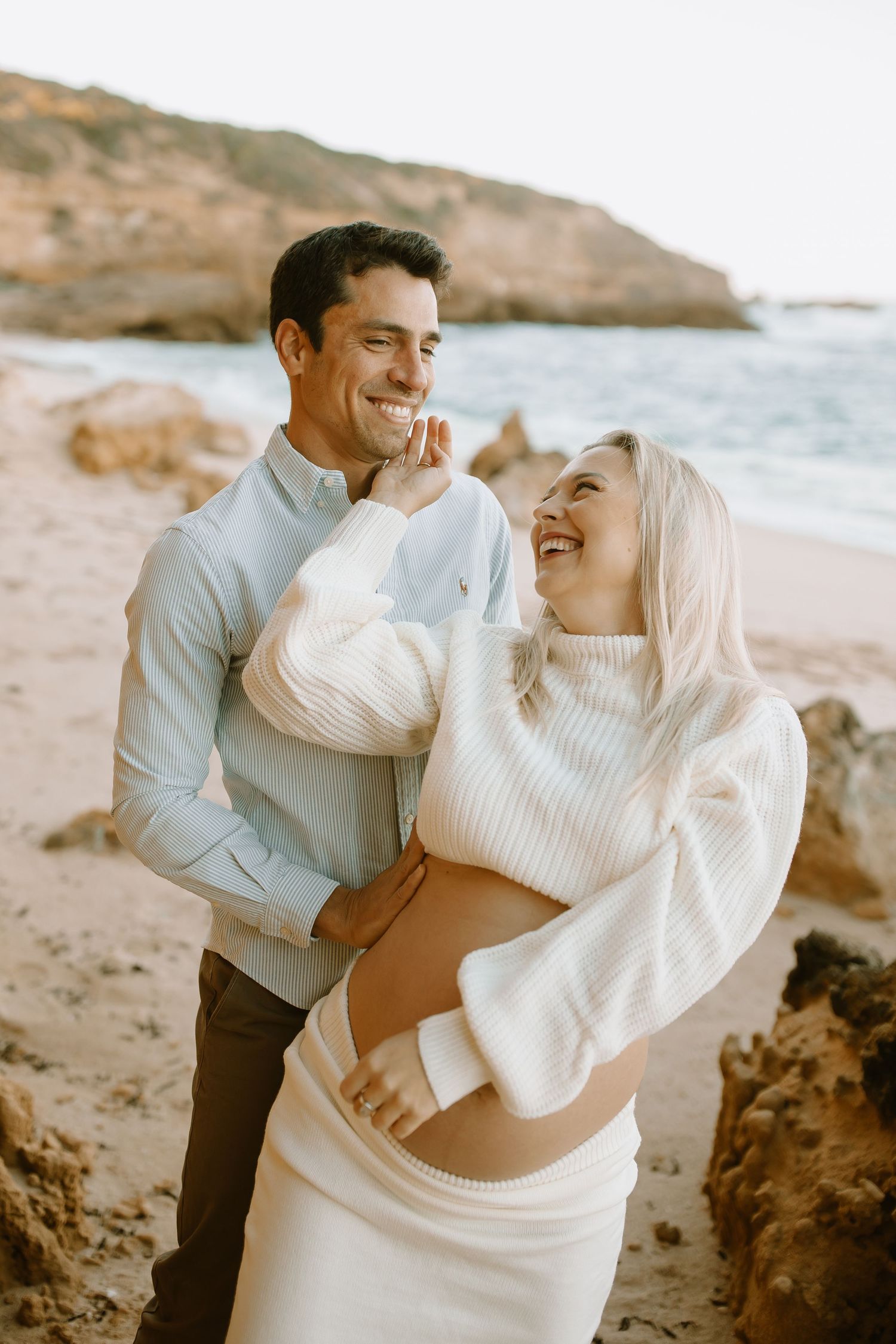 A couple in white sweaters shares a joyful moment together on a rocky beach during golden hour.
