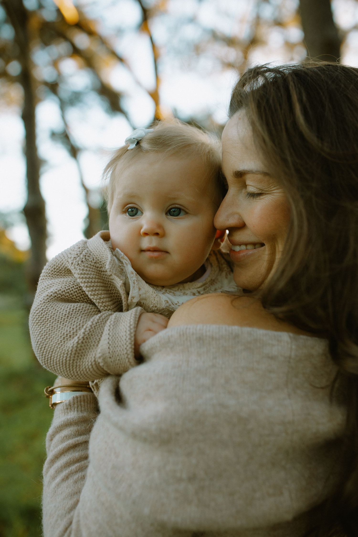 A parent in a beige sweater shares an intimate moment with their baby against a soft-focused natural backdrop.
