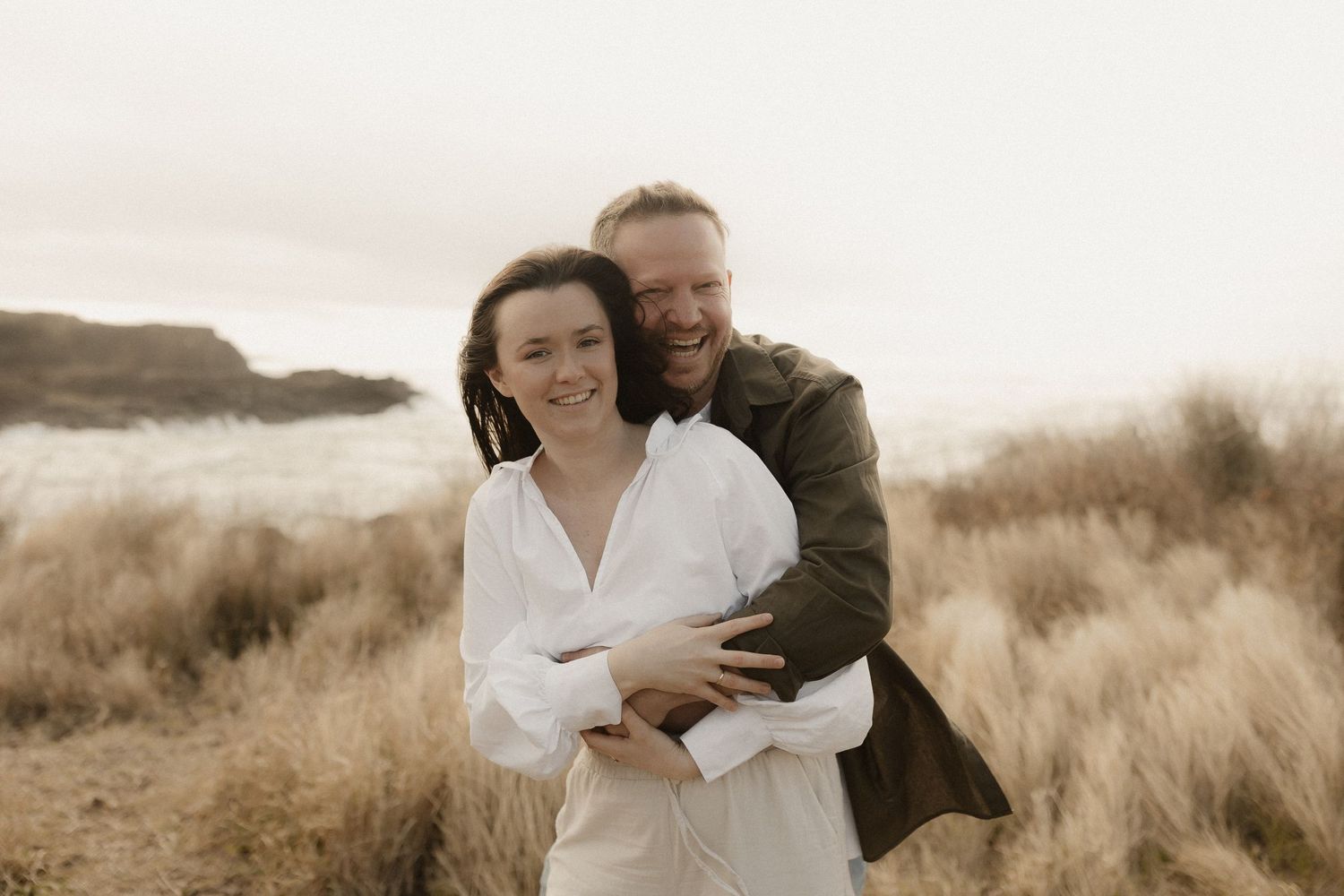 Romantic couple embraces in sepia tones against a backdrop of windswept beach grass at sunset.