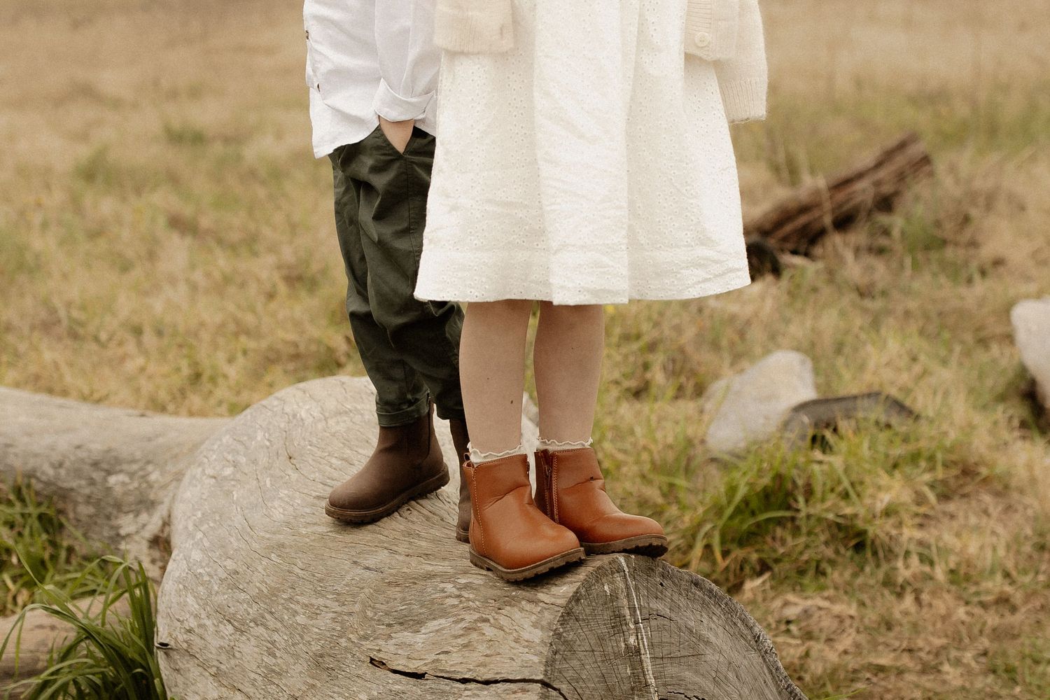 Woman in white dress and brown leather boots stands in grassy field with rustic wooden elements.