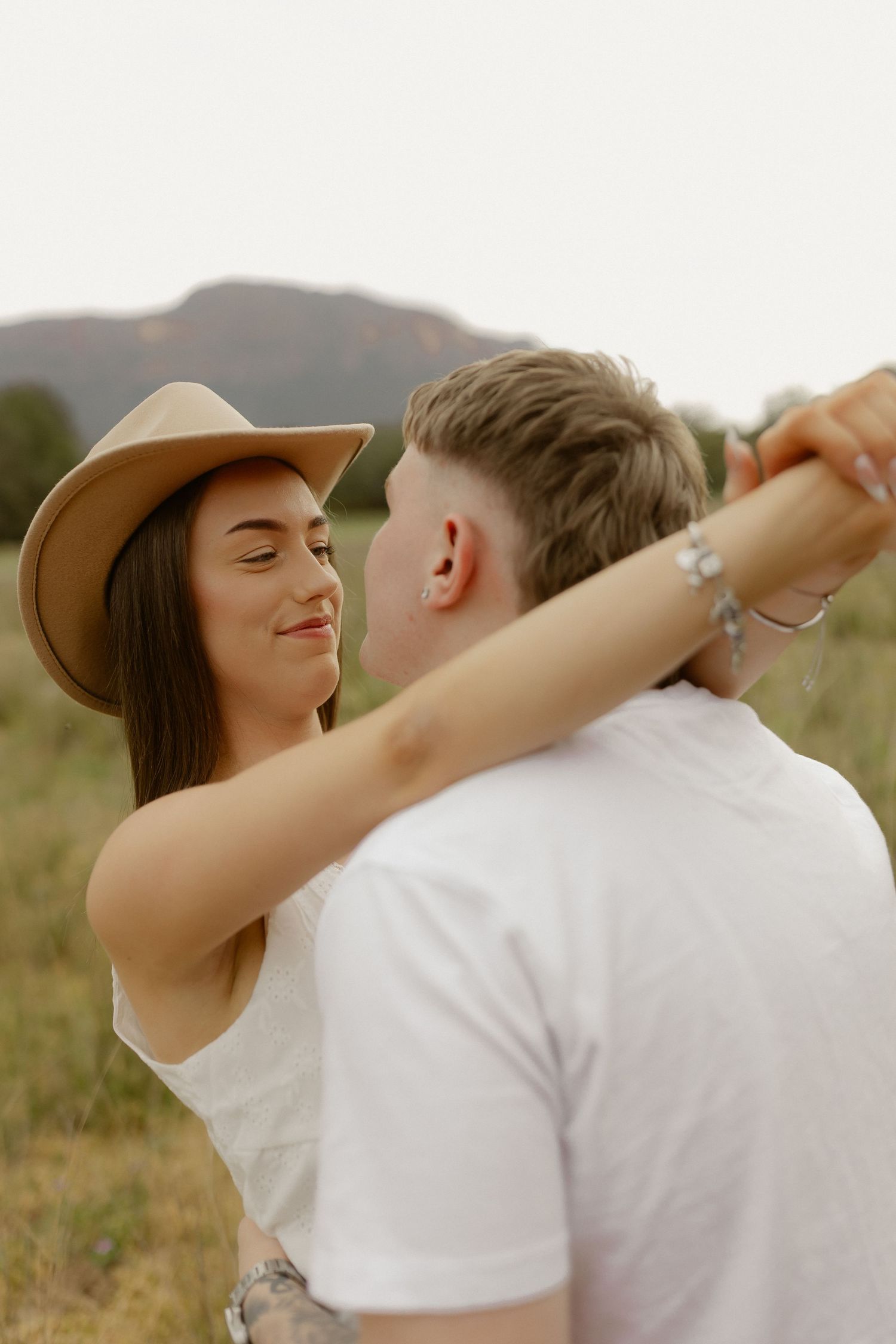 A romantic couple embraces in a field at sunset with mountains in the background.