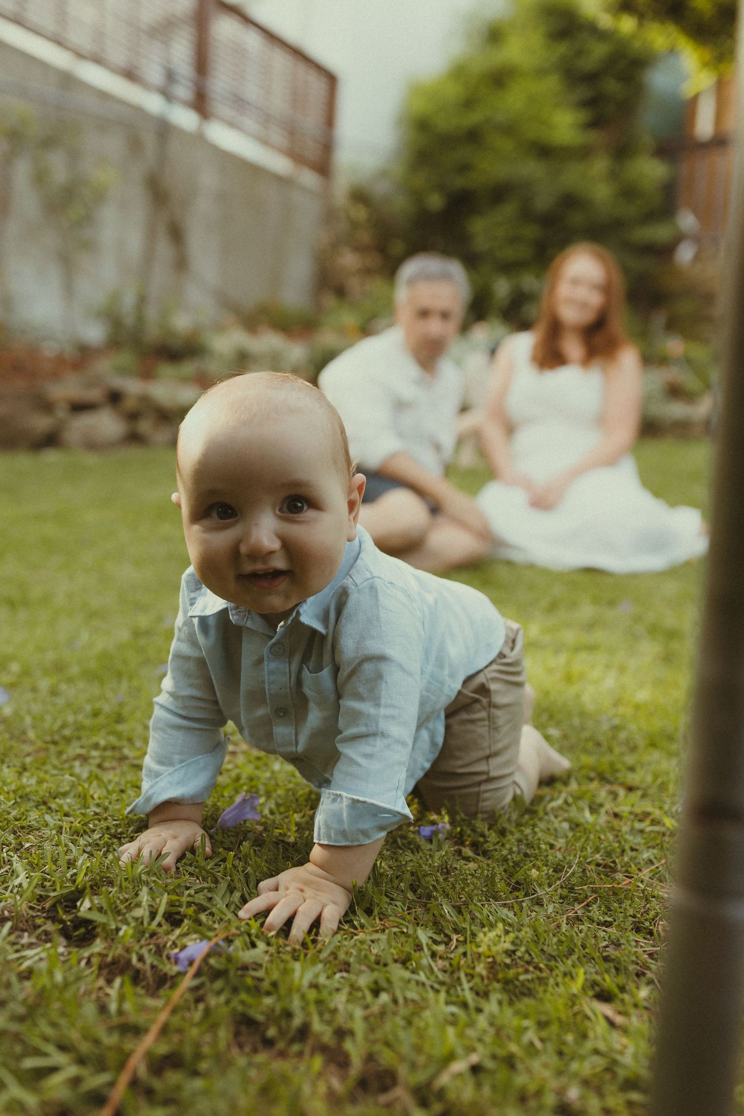 A baby in a light blue shirt crawls on green grass during an outdoor photo session.