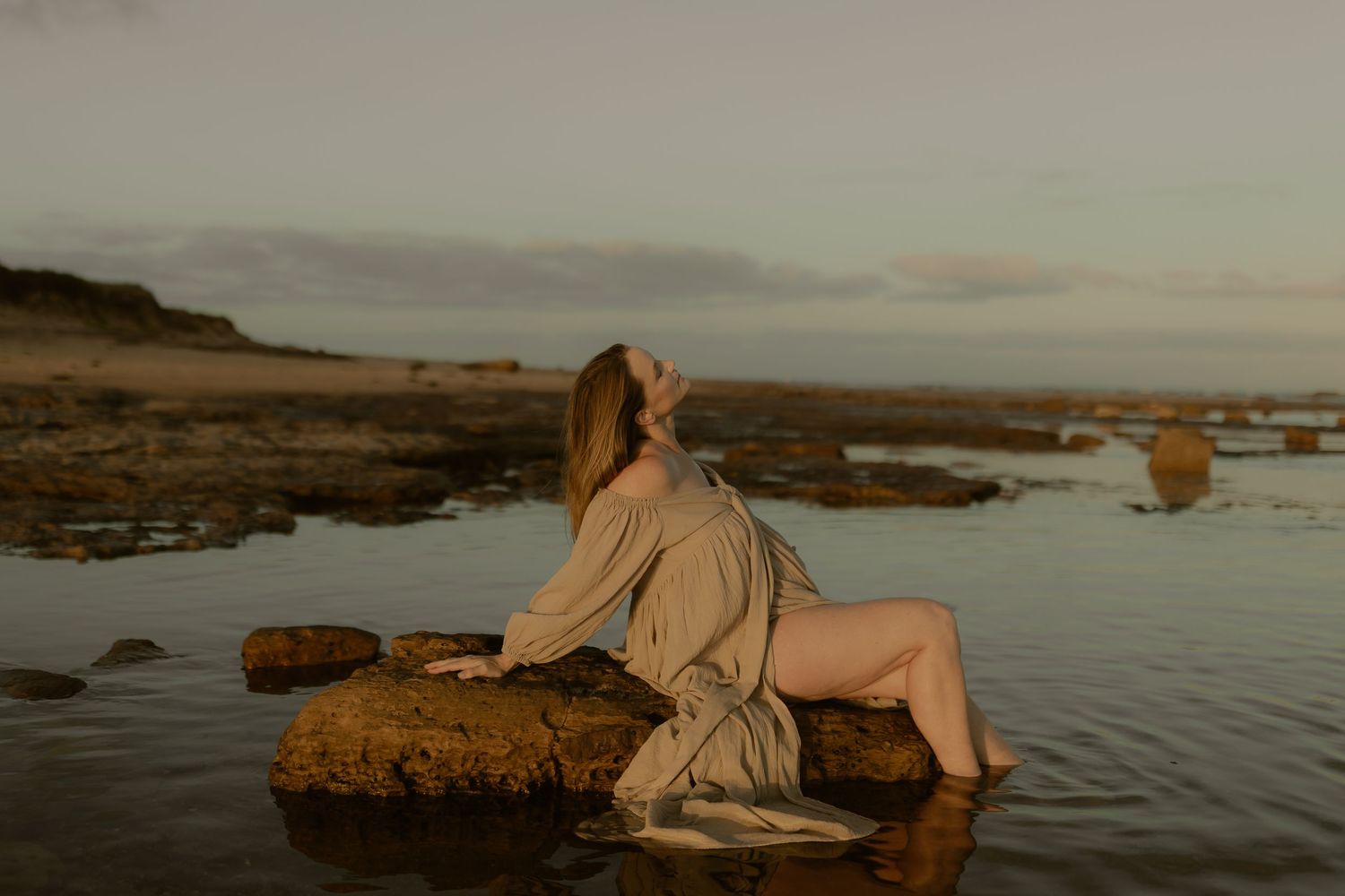 A moody sunset photograph of someone reclining on coastal rocks with water surrounding them.