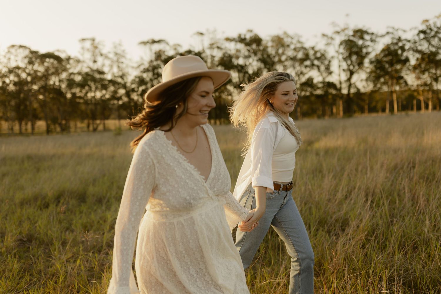 Two friends walking through a sun-drenched field at golden hour wearing white dresses and casual attire.